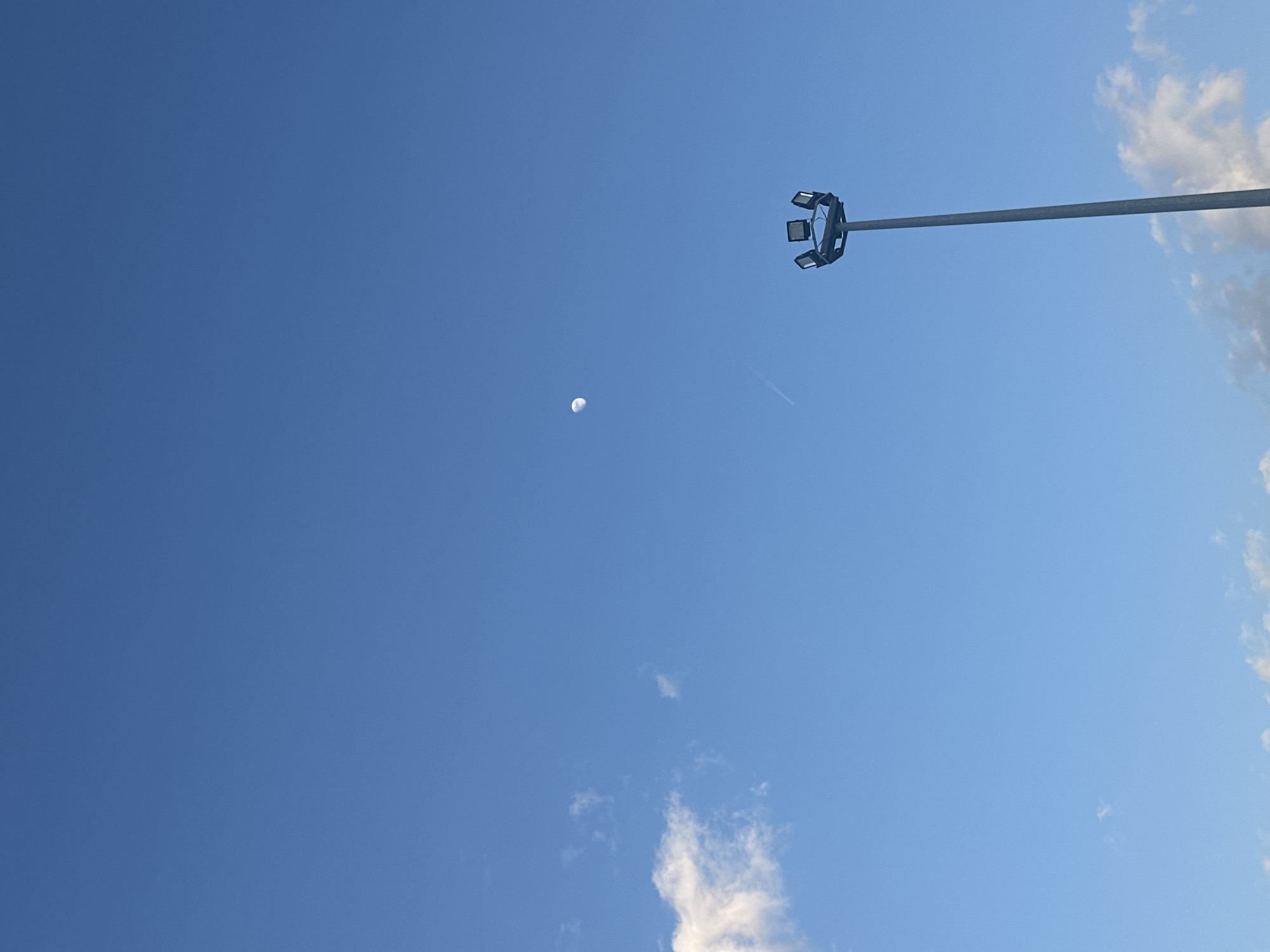 The image depicts a daytime sky in Manisa, Turkey. A light blue sky is visible, with a crescent moon towards the center-left. A light pole with multiple spotlights extends from the upper right corner, pointing towards the center of the frame. Wispy clouds are present along the right edge of the image and toward the bottom. A contrail from an aircraft is visible near the moon.