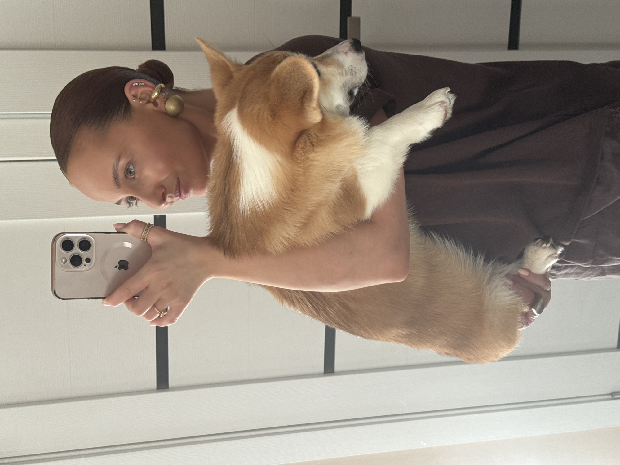 A fair-skinned woman with brown hair in a bun and multiple ear piercings is holding a tan and white corgi dog in her arms and taking a photo of them both with a silver iPhone. She is wearing a brown shirt. The background is a white door with horizontal lines and a black vertical line. The location in the city of Chaikovsky, Russia, is not discernable from the image.