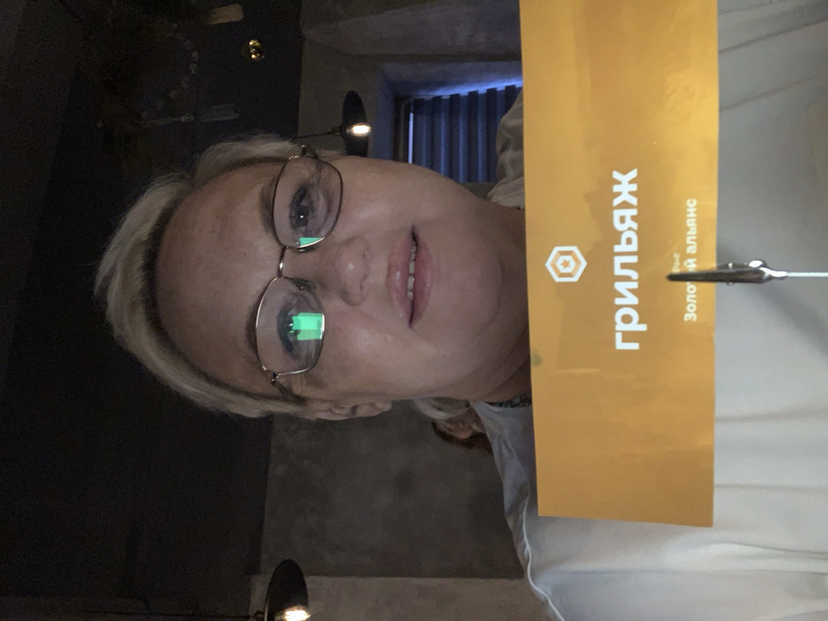 An indoor, close-up, chest-up photograph depicts a woman with short, light-colored hair and gold-framed eyeglasses, looking directly at the camera with a slight smile. A green rectangular light source is reflected in her left eyeglass lens. She is wearing a light-colored top. In the foreground, she holds a horizontal orange sign that displays "грильяж" (grilyazh) in prominent white Cyrillic text, with "Золотой альянс" (Golden Alliance) written below it in smaller text. A metallic clip secures the bottom edge of the sign. The background shows a dimly lit interior, characteristic of a cafe or restaurant located in Chaikovsky, Russia. Features include dark, textured walls, two industrial-style pendant light fixtures with exposed bulbs, and a horizontal blind visible through an opening on the right. A blurry, indistinct figure is partially visible in the deep background to the left of the woman's head.