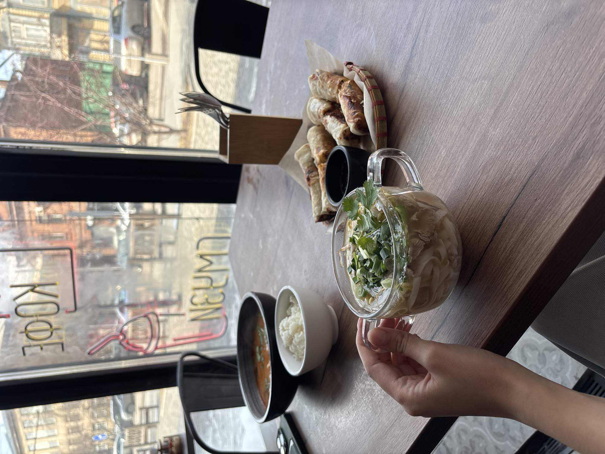 The image displays an indoor dining scene at a cafe or restaurant, with a focus on a table set next to a large window. A person's hand is visible, holding a clear glass mug filled with a noodle soup, likely pho, garnished with green onions and other herbs. On the light-colored wooden table, there's also a black bowl containing a reddish soup (possibly tom yum), and a small white bowl with white rice. A rustic basket holds several spring rolls or fried wraps, positioned next to a small black cup. Cutlery is arranged in a wooden holder on the table.
Through the window, a street scene is visible, showing buildings, a parked car, and trees with bare branches, suggesting a cooler time of year. The natural light indicates it is daytime. Reflected on the window glass is a neon sign displaying Russian text, which appears mirrored from this perspective. The readable text includes "КОФЕ" (Coffee) and "НЕЙМЕД" (Named), along with a stylized graphic that resembles an electrical plug. This scene captures a meal in a public establishment, possibly in Krasnoyarsk, Russia, given the Russian text.