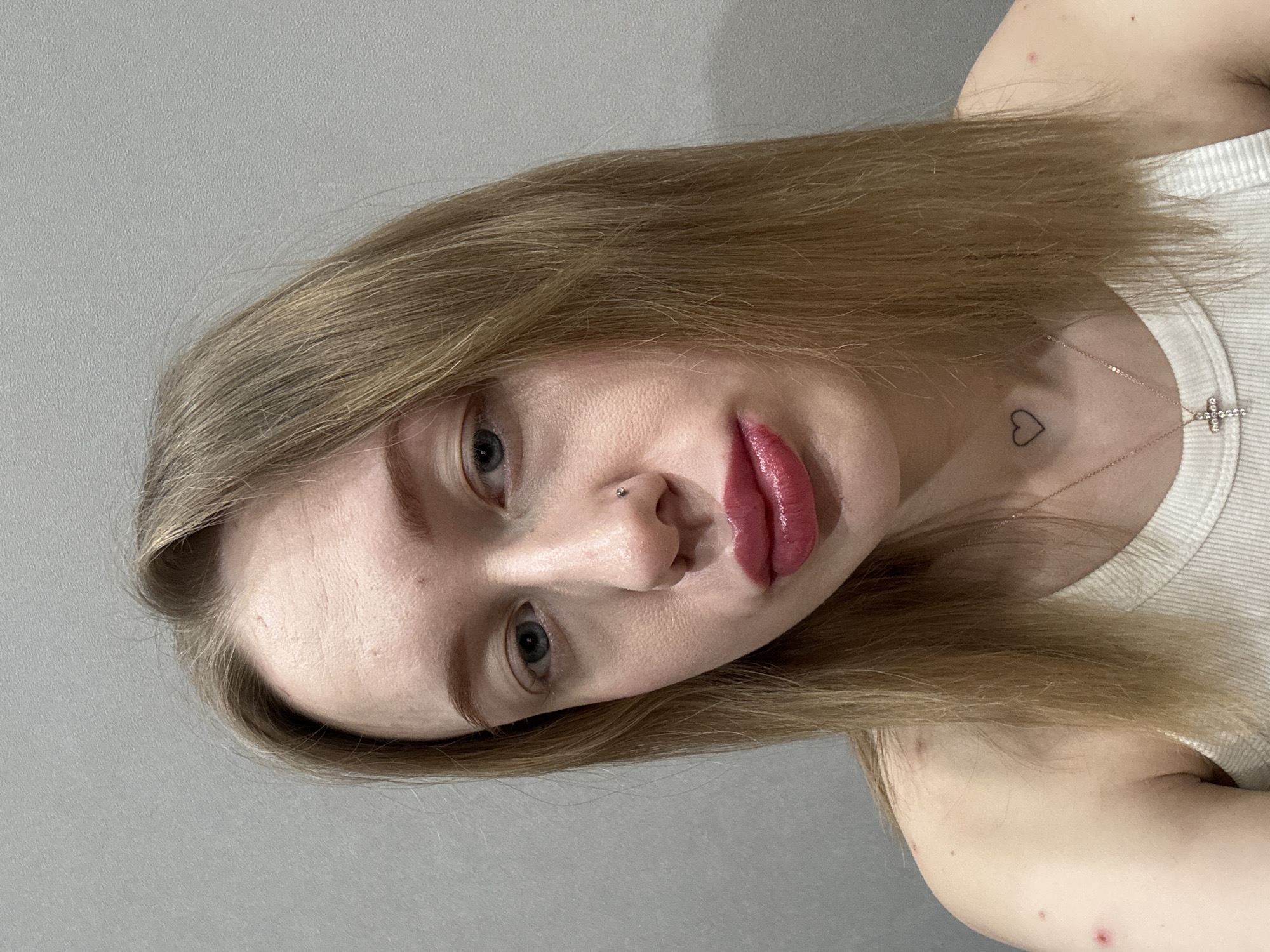 A young adult female is depicted in a close-up, chest-up portrait against a plain, light grey, textured wall. She possesses long, straight, blonde hair and light-colored eyes. A small silver stud piercing is visible in her left nostril, and her lips are coated with glossy dark pink lipstick, appearing slightly pursed. A small, black outline heart tattoo is present on her anterior neck, slightly left of the midline, above a thin gold chain necklace with a small, clear gemstone-studded cross pendant. She is wearing a white ribbed tank top. A distinct red lesion is observed on her left upper arm. The subject is directly facing the camera. This scene is located in Rostov-on-Don, Russia.