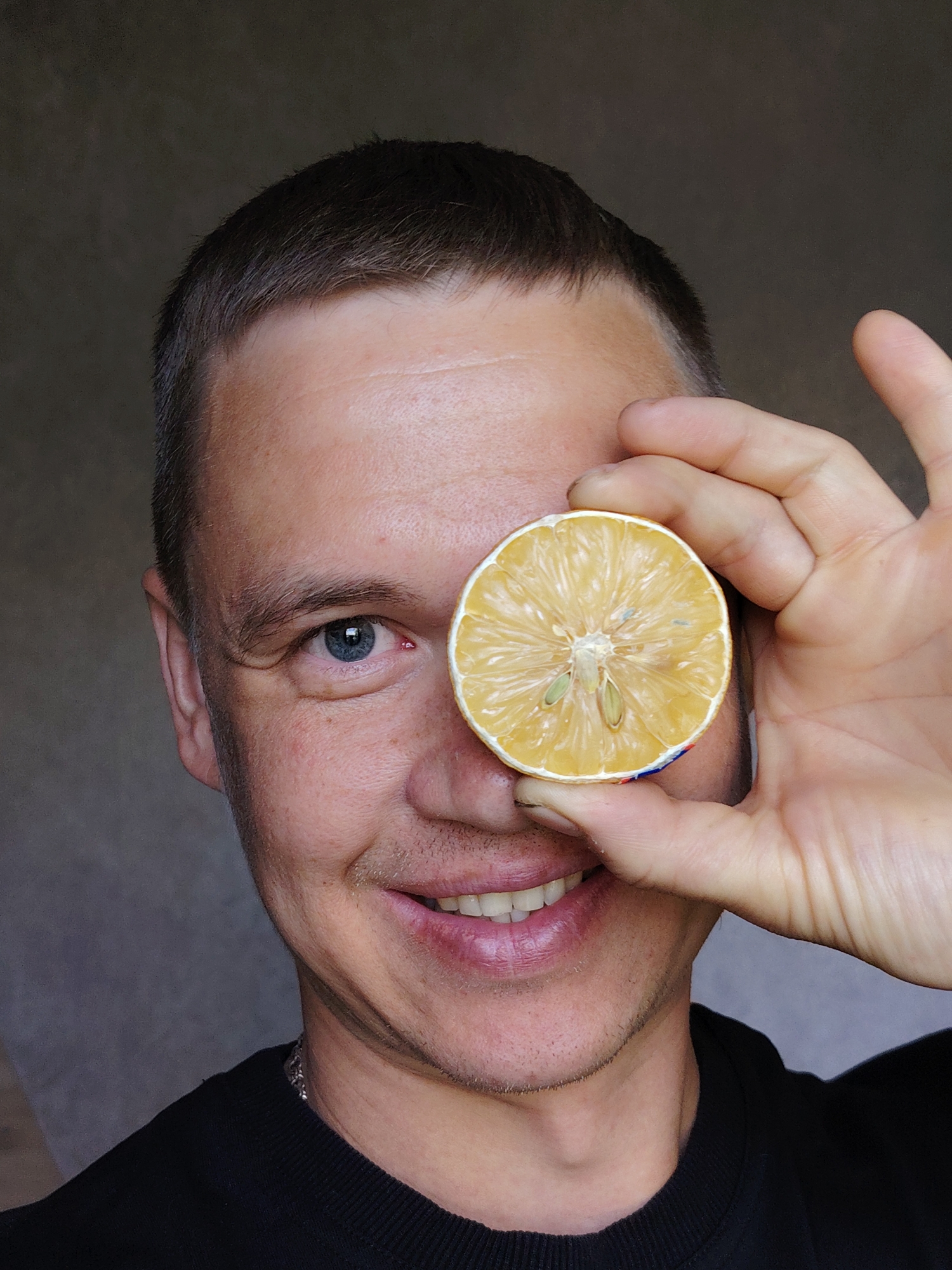 A close-up shot shows a man with short, dark hair smiling and holding a slice of dried lemon in front of his face, obscuring one eye. His skin has fair tones. The eye that is visible is blue. He is wearing a black crew neck shirt. The background is a muted gray. The image was likely taken indoors. There is no visual evidence to confirm the location is in Chaikovsky, Russia.