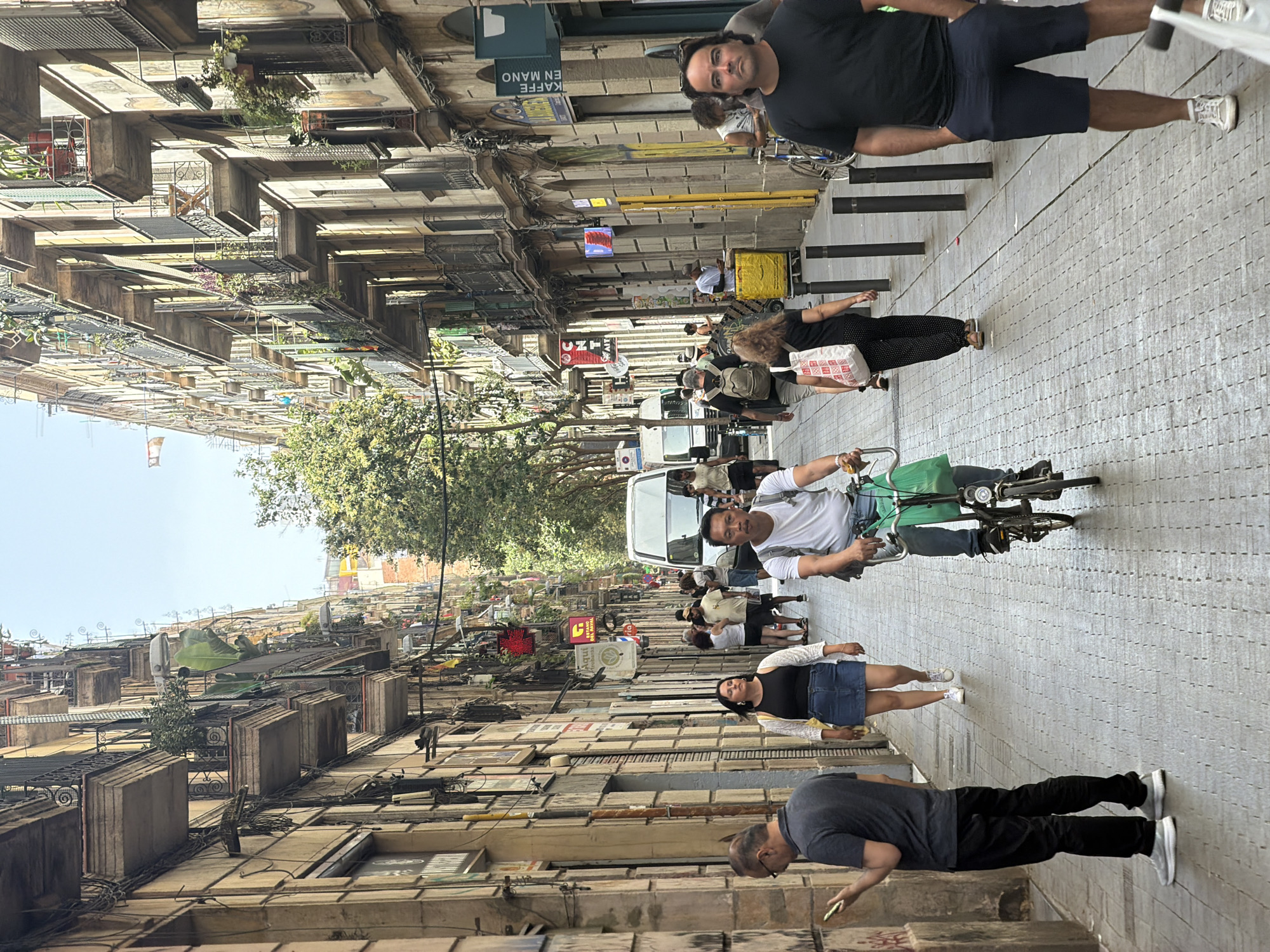This media file depicts a street scene in Barcelona, Spain. The perspective is from a high vantage point looking down a narrow street lined with multi-story buildings. Several people are visible, some walking, others on bicycles, and one person appears to be pushing a bicycle with a green bag attached. One individual is dressed in a dark t-shirt and shorts, while another wears a black t-shirt and a denim skirt. A person in the foreground is wearing a dark t-shirt and black pants.
The street appears to be paved with a light-colored, textured material. Sunlight is casting shadows, suggesting it is daytime. The architecture of the buildings suggests an urban environment, with balconies and signs visible. Some of the signs are in Spanish, with one clearly reading "KAFFE EN MANO" which translates to "Coffee in Hand." Another sign is red and has a cross symbol, possibly indicating a pharmacy or clinic. The overall mood seems to be that of everyday street activity. The orientation of the image is such that the people and buildings are mostly vertical, indicating the original capture was likely in portrait mode, but the provided media is rotated.