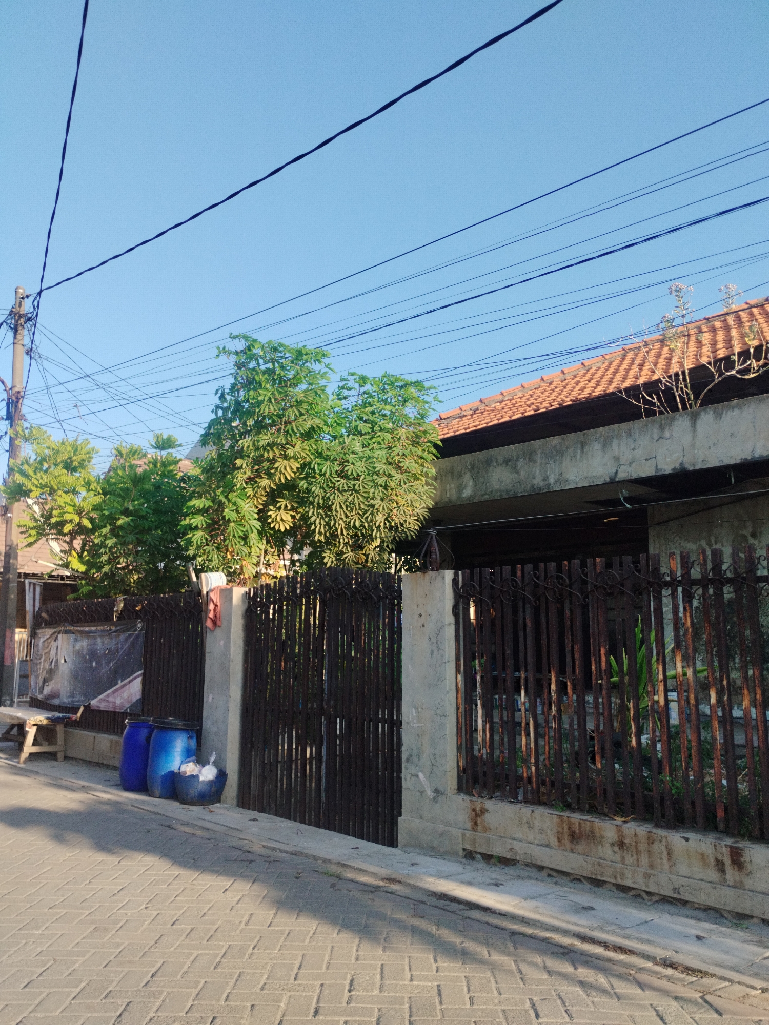 The image depicts a street scene in Surabaya, Indonesia. In the foreground, a paved sidewalk made of light gray bricks runs diagonally from the bottom left to the top right. A dark shadow, cast by an unseen object, stretches across the sidewalk. To the left of the sidewalk, there are two large blue plastic barrels and a smaller dark blue bin containing white plastic bags. A wooden bench with a dark seat is positioned behind the barrels. A section of a wall, possibly concrete or plaster, is visible to the right of the bench. Further back, a dark metal fence with vertical bars and decorative elements runs along the edge of a property. Behind the fence, a low building with a concrete facade and a terracotta tiled roof is visible. Lush green trees with dense foliage are growing in front of and over the fence. Overhead, multiple dark electrical wires crisscross against a clear, pale blue sky, with a utility pole visible on the left side of the image. The overall impression is a snapshot of a residential or commercial area in an Indonesian city, characterized by its tropical vegetation and infrastructure.