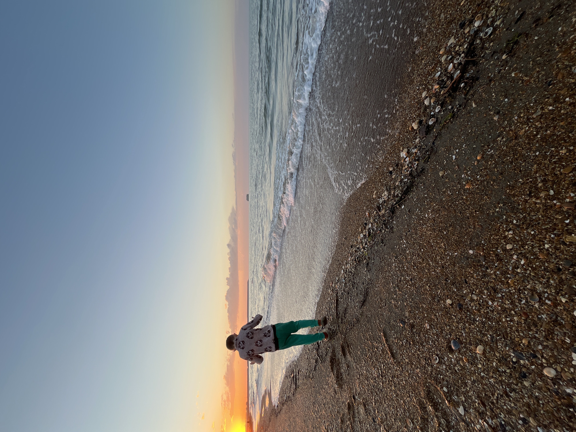 A young boy stands on the beach at sunset in Kaspiysk, Russia. He is wearing green pants and a light-colored sweatshirt with a repeating pattern. The beach is composed of small pebbles and shells. The water is shallow, and small waves are visible. The sky is a gradient of blue, orange, and pink.