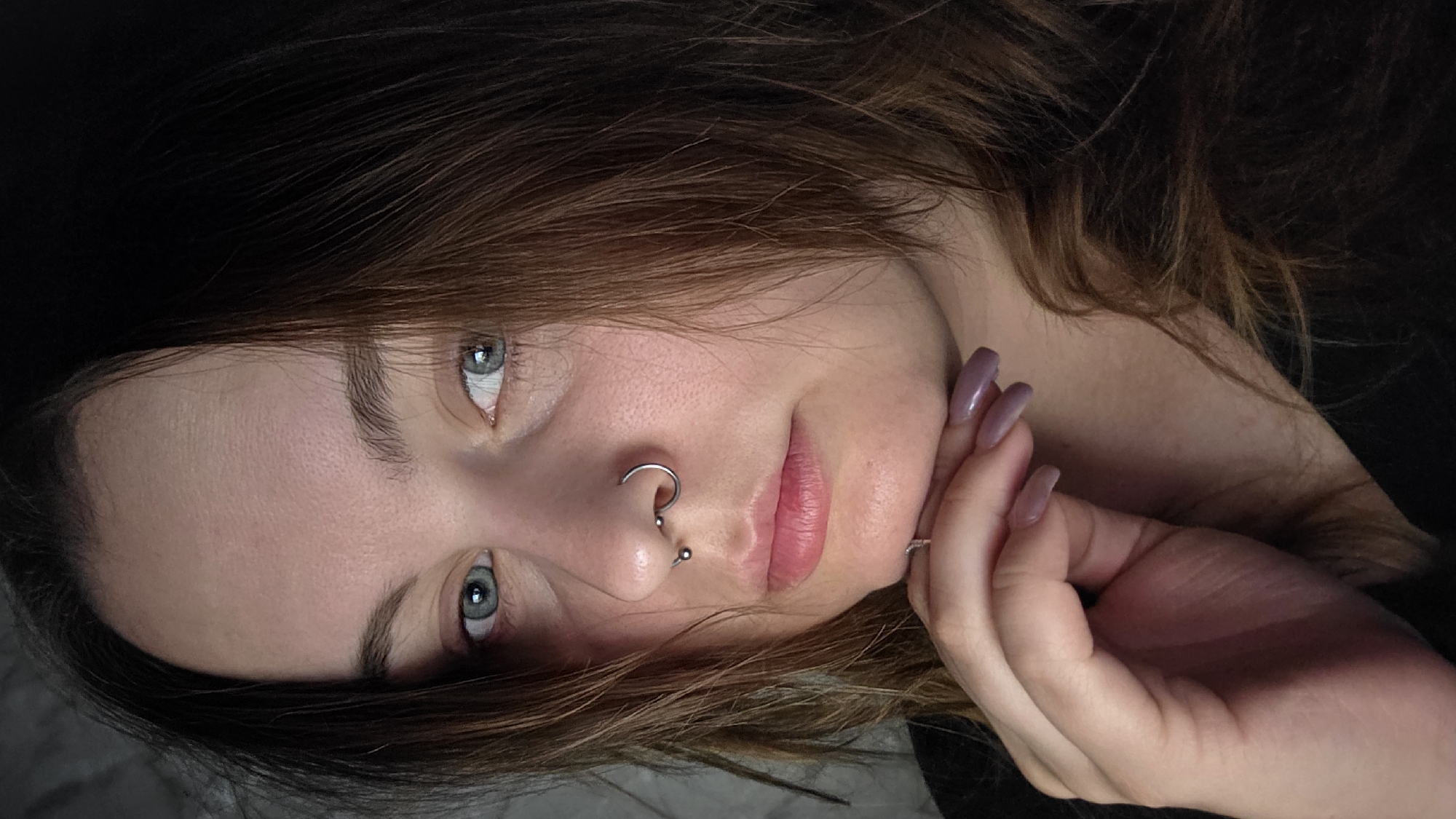 A close-up portrait captures a woman with fair skin, light brown hair, and blue eyes. She has a nose ring and a small hoop piercing. Her fingernails are painted a mauve color. The background is dark and indistinct. The image is rotated counterclockwise.