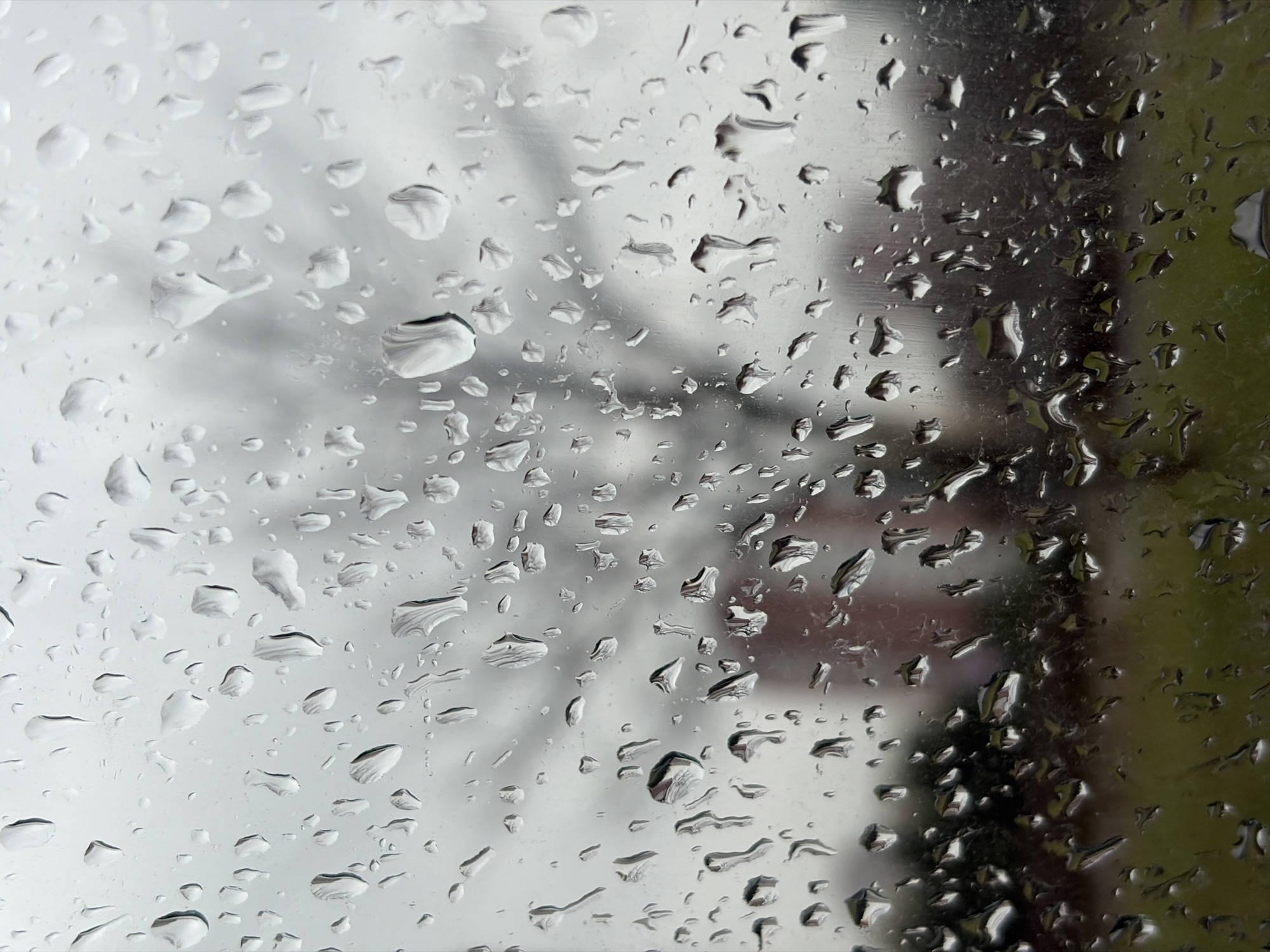 The image shows a close-up view of water droplets on a windowpane. Through the window, an out-of-focus background reveals a gray sky, possibly indicating overcast weather, along with blurred shapes suggesting buildings or vegetation. The droplets themselves vary in size and shape, refracting the light and distorting the background. The lower right portion of the window reflects a dark green mass. There are no people, subjects or actions.
