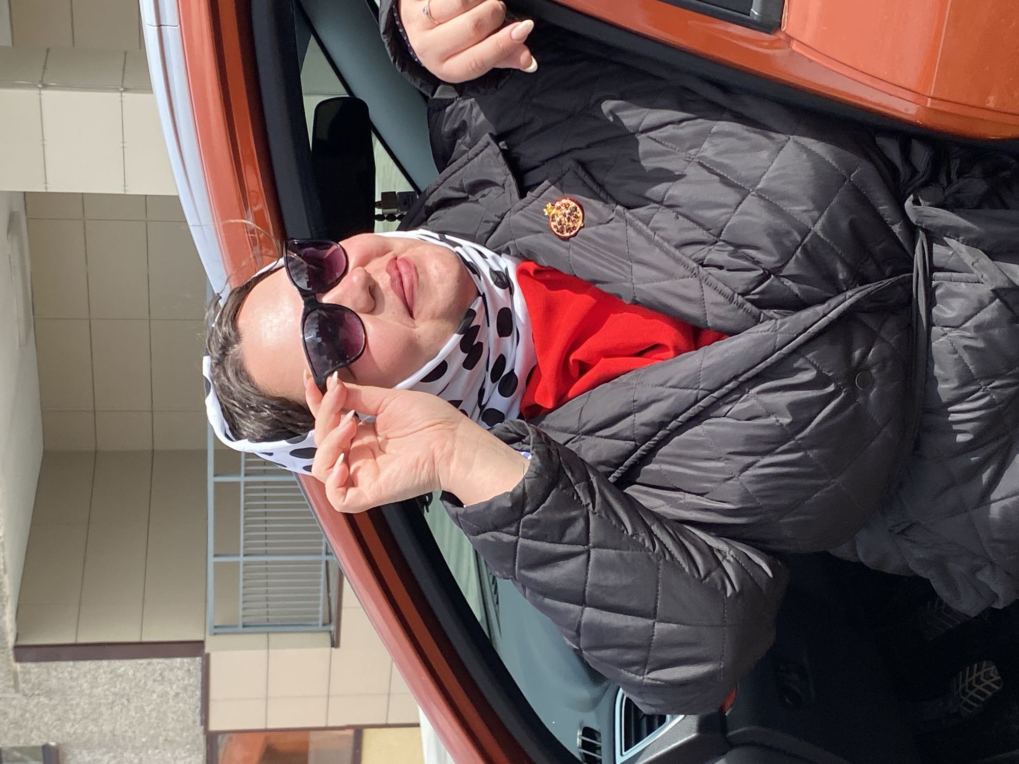 A woman is seen from the chest up, positioned inside an orange car with a black interior. The car appears to be stationary. The woman is wearing a black quilted jacket with a gold brooch on the lapel, a red garment underneath, and a white scarf with black polka dots tied around her head. She is also wearing dark sunglasses and has manicured nails. She is holding the sunglasses in her right hand, adjusting them. The background shows parts of a building with windows and light-colored exterior walls. The photo was taken outdoors in daylight. The location is Марковский, Russia.