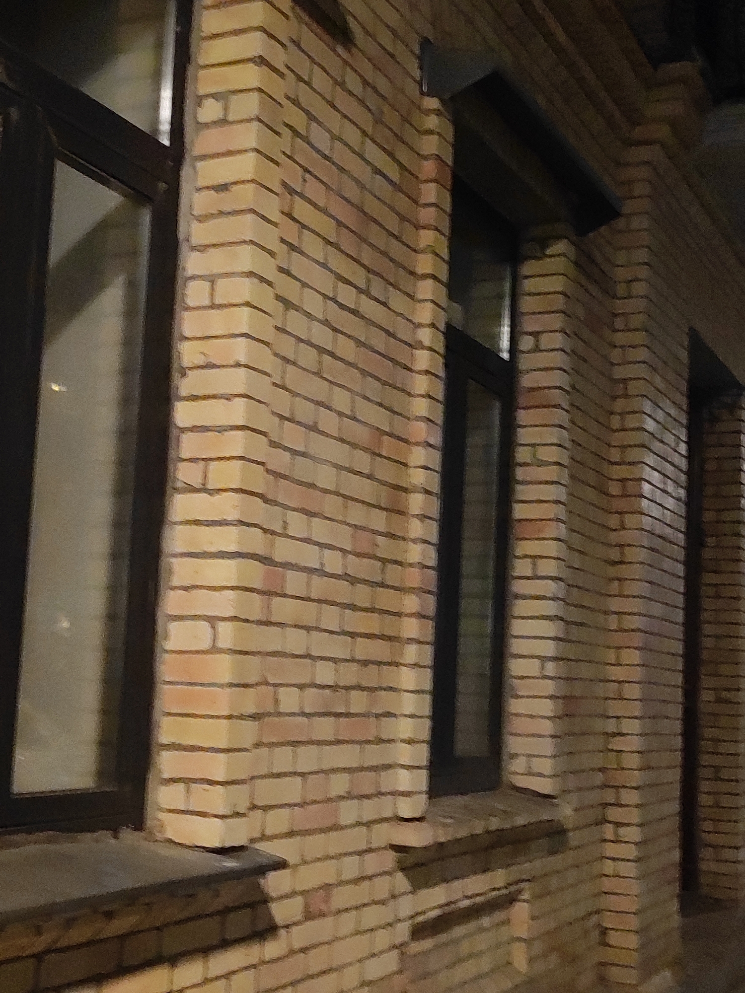 The image shows a close-up view of a light brown brick building exterior at night. The building has dark framed windows and a dark colored awning above one of the windows. The bricks are laid in a horizontal pattern with visible mortar lines. The lighting suggests artificial light sources are present, casting shadows on the wall and window sills. The overall scene is devoid of people or any discernible activity. The image is a detail of an urban environment, likely in Batumi, Georgia, given the provided context.