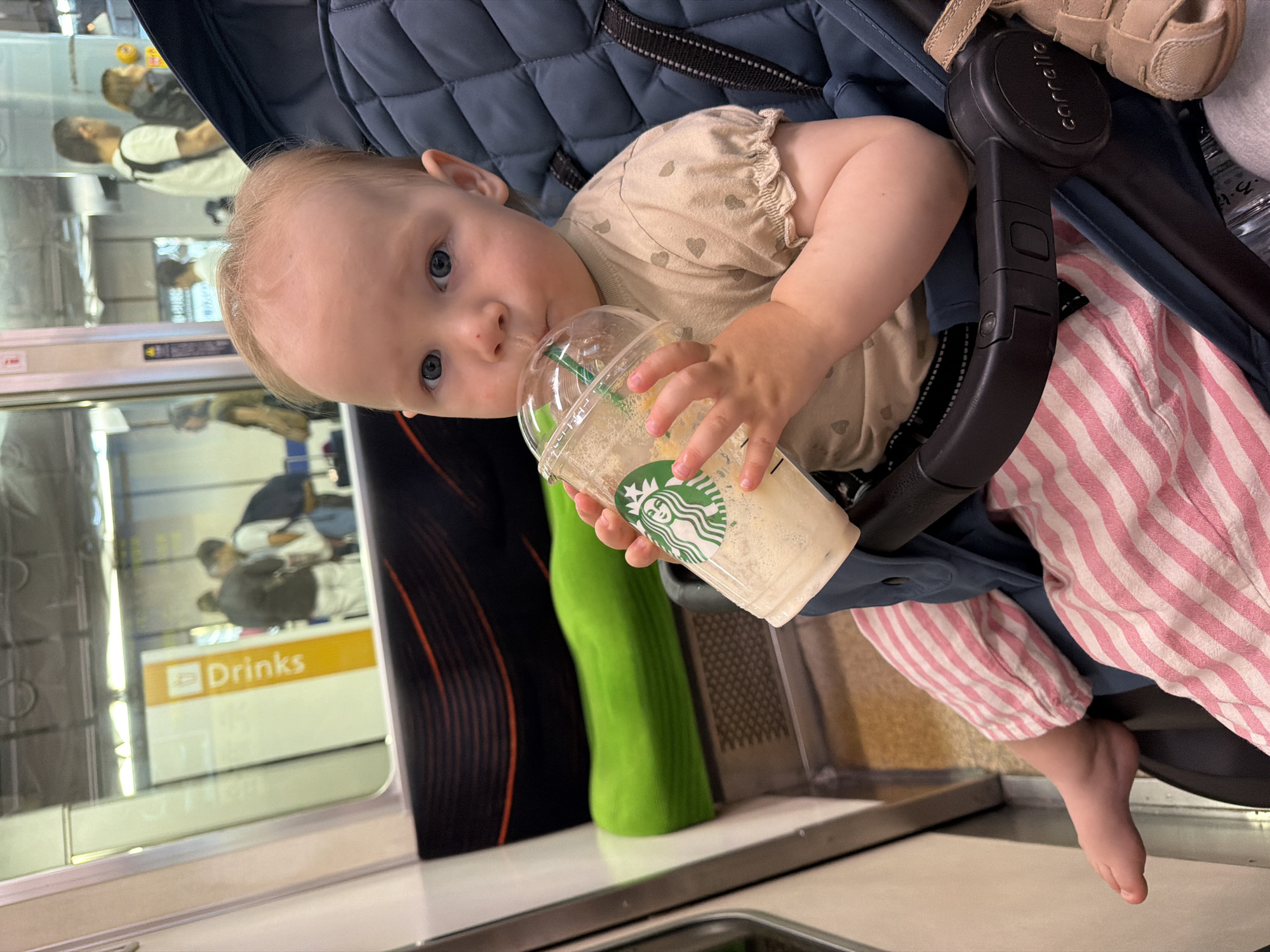 A baby with light hair and blue eyes is seated in a dark blue "Carrello" brand stroller inside a train carriage, looking directly at the camera. The child is wearing a light beige t-shirt with faint heart patterns and pink and white striped pants, and is drinking from a clear plastic Starbucks cup with a green straw, containing a light-colored blended beverage. A single light brown sandal is visible on the right side of the stroller. The train interior features a distinctive lime green, horizontally ribbed bench seat to the left of the stroller, with a dark patterned wall above it. Through the train window, reflections show multiple blurry adult figures and a vertical orange sign with the word "Drinks" in white, suggesting a train station platform. The train's interior design, particularly the lime green seating, is characteristic of the Yamanote Line in Tokyo, Japan.