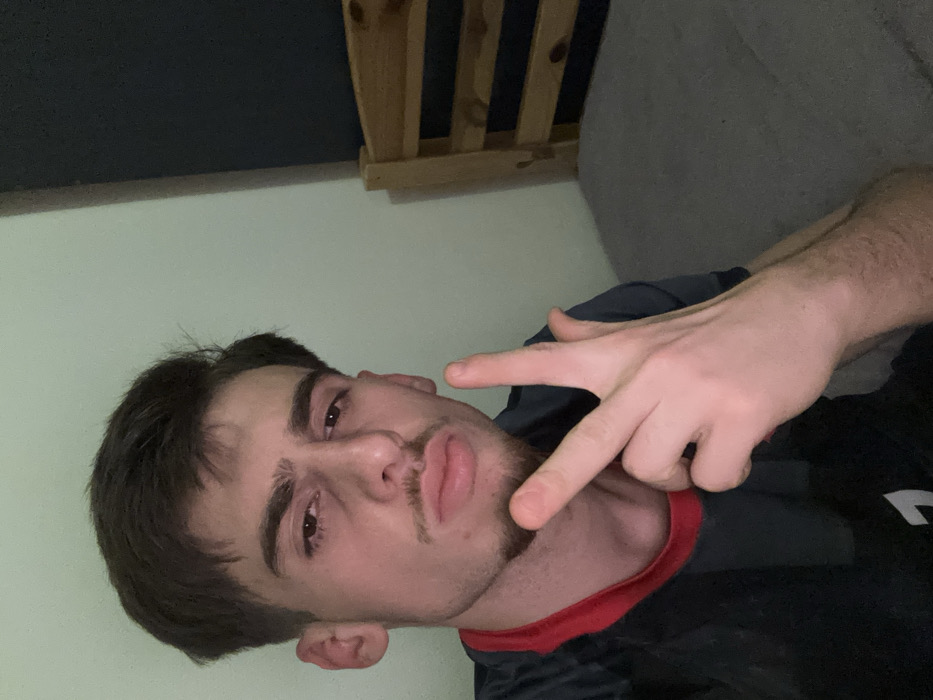 A selfie shows a young man with dark hair, brown eyes, and light skin. He has a mustache and a goatee. He's wearing a dark shirt with red trim around the neck. He's holding his hand up, making a gesture with his index and middle fingers. The background includes a light-colored wall and what appears to be a wooden structure or headboard above and to the side. Part of a gray cushion is visible behind his shoulder. The image is rotated sideways.