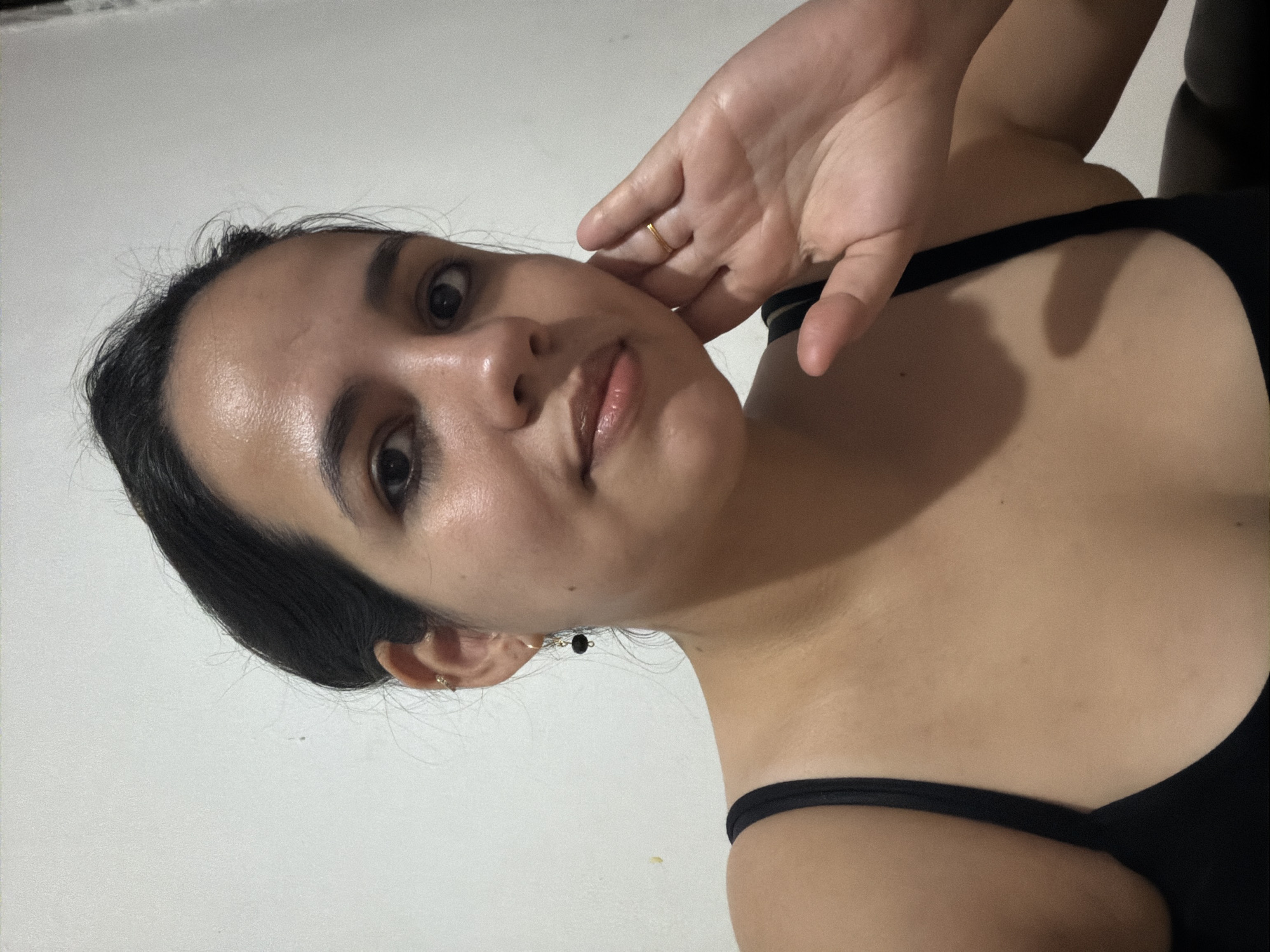 The image displays an adult female from the chest up, positioned indoors in Ciudad Guayana, Venezuela. She faces the viewer directly with a neutral expression. Her hair is dark and pulled back, and her eyes show dark eyeliner on the lower lash line. She wears a black tank top. Her visible left ear is adorned with a small gold-colored hoop piercing in the upper helix and a dangling earring featuring a black spherical bead. Her right hand is raised, with the palm forward and fingertips lightly contacting her right cheek. A thin gold-colored ring is visible on her right ring finger. The background behind the subject is a plain, light-colored wall with minor textural variations.