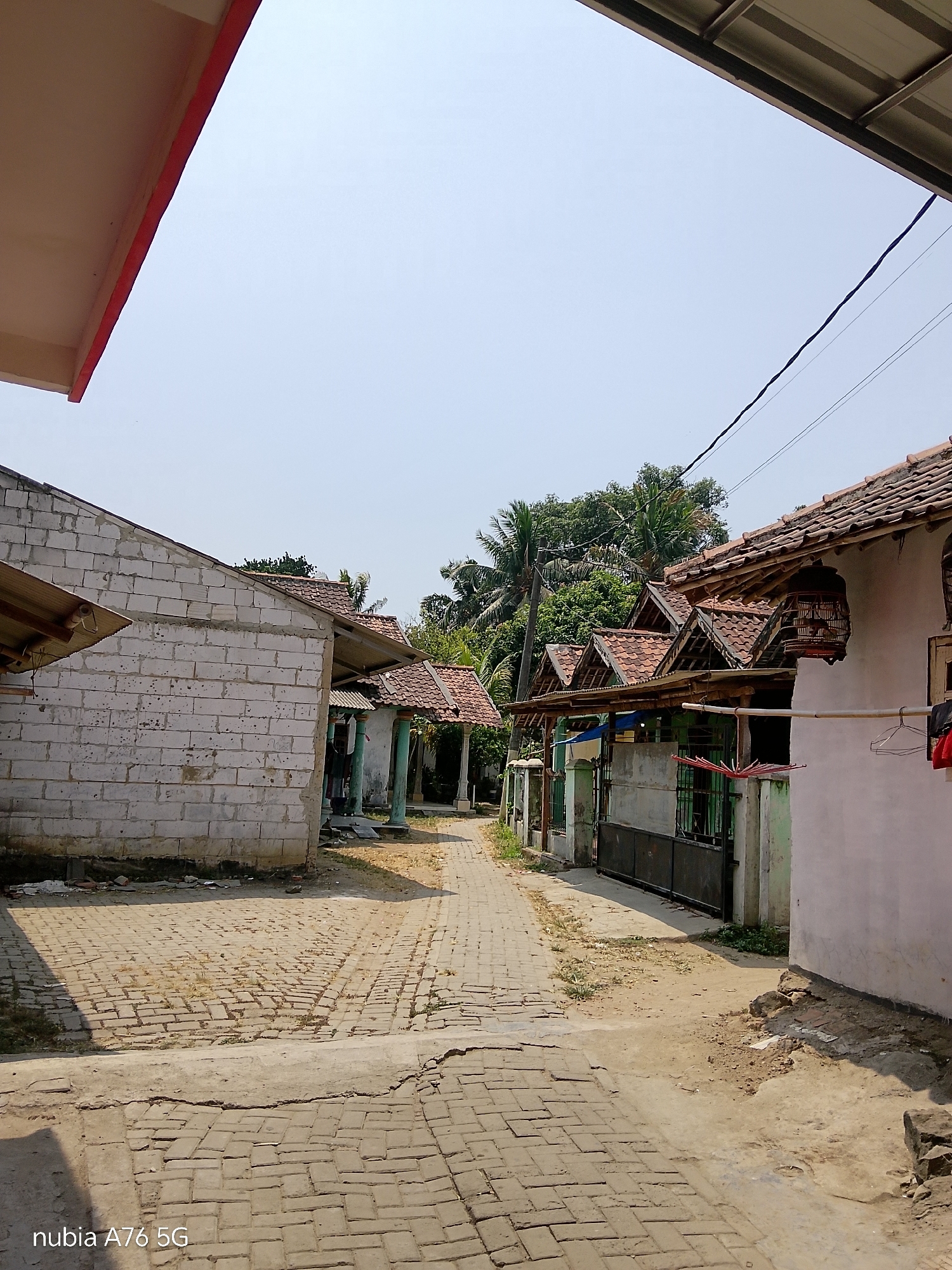 The image shows a narrow, paved alleyway in a residential area in Cakung, Indonesia. The alley is lined with simple, single-story houses with tiled roofs. The buildings are constructed with a mix of materials, including brick and concrete blocks. Some houses appear to be under construction or renovation, with exposed brickwork visible on one building. The alleyway is partly shaded by the roofs of the houses and by trees in the background. A clothesline with items hanging on it is visible on the right side of the alley. The sky is clear and bright. The overall impression is one of a humble, everyday scene in a rural or semi-urban Indonesian setting.