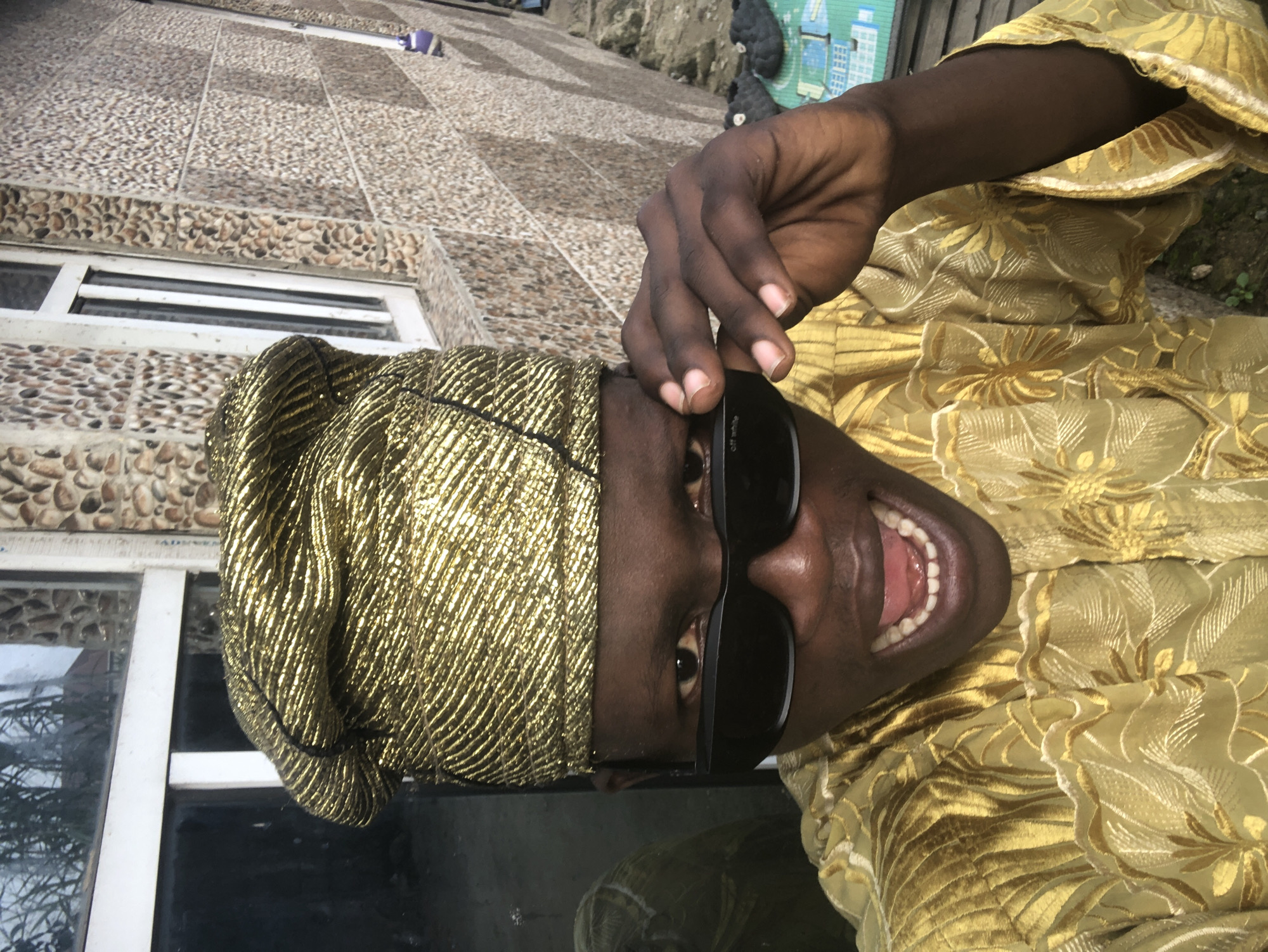 A young Black man in Ado, Nigeria, poses for a selfie outdoors, wearing a traditional gold patterned cap and matching garment. He is holding his dark-rimmed sunglasses with his right hand, his mouth open in a wide smile, revealing his teeth. The background features a textured wall made of small, multi-colored stones and a white-framed window. A reflection of the man and the environment can be seen in the window glass. The ground in front of him has some scattered objects, including what appears to be a green mat with a grid pattern and some dark-colored footwear. The overall lighting suggests it is daytime.