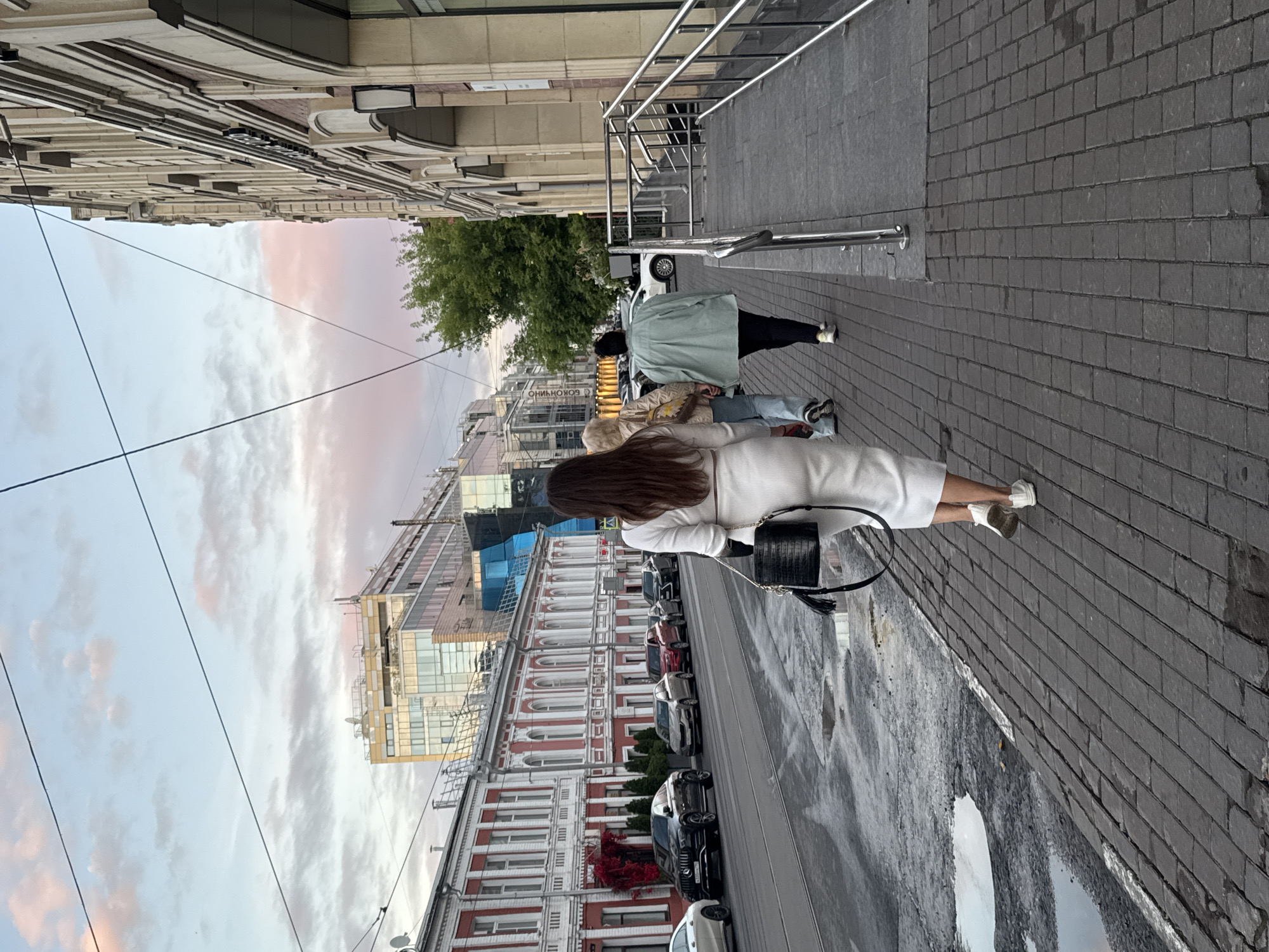 The image shows a street scene in Nizhny Novgorod, Russia, featuring several people walking on a sidewalk alongside a road. The architecture suggests an urban environment with a mix of older, classical buildings with red and white facades, and more modern glass-fronted structures.
In the foreground, a woman in a white dress and white sneakers is walking away from the camera, carrying a black textured handbag with a tassel. Behind her, two other individuals are walking in the same direction; one appears to be wearing a light-colored jacket and jeans, and the other a light green oversized jacket and dark pants.
The street in front of the buildings is wet, with several puddles visible, indicating recent rain or a wet surface. Several cars are parked along the street. The sky above is a mix of blue and soft pink clouds, suggesting it is either early morning or late evening. Overhead, power lines are visible against the sky. The sidewalk is paved with dark rectangular cobblestones. A metal railing and ramp structure are visible to the right of the pedestrians, leading towards a building.