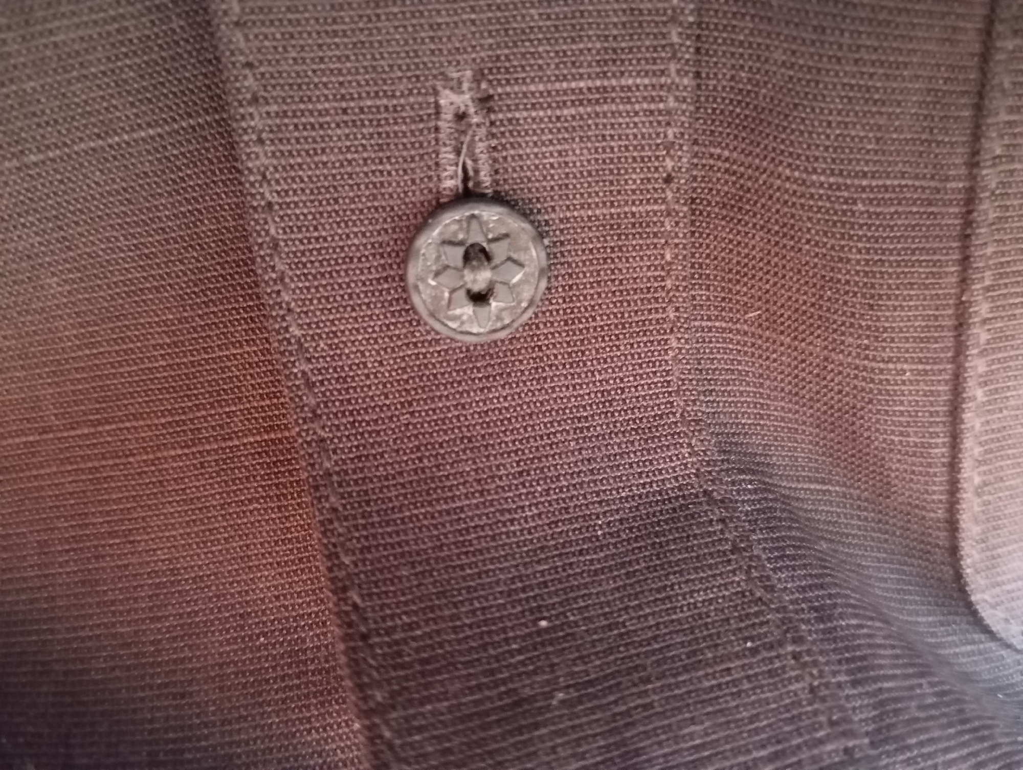 A close-up, eye-level, medium shot shows a single, grey, round button with a starburst pattern in its center. The button is attached to a dark brown fabric with a subtle ribbed texture, likely from a piece of clothing such as a jacket or shirt. The fabric appears to be made of a woven material. The lighting creates a gradient of light and shadow across the fabric, with the left side appearing lighter and redder, and the right side darker and more subdued. The image is highly detailed, focusing on the texture of the fabric and the design of the button. No specific location in the city of Unknown, Unknown is depicted.