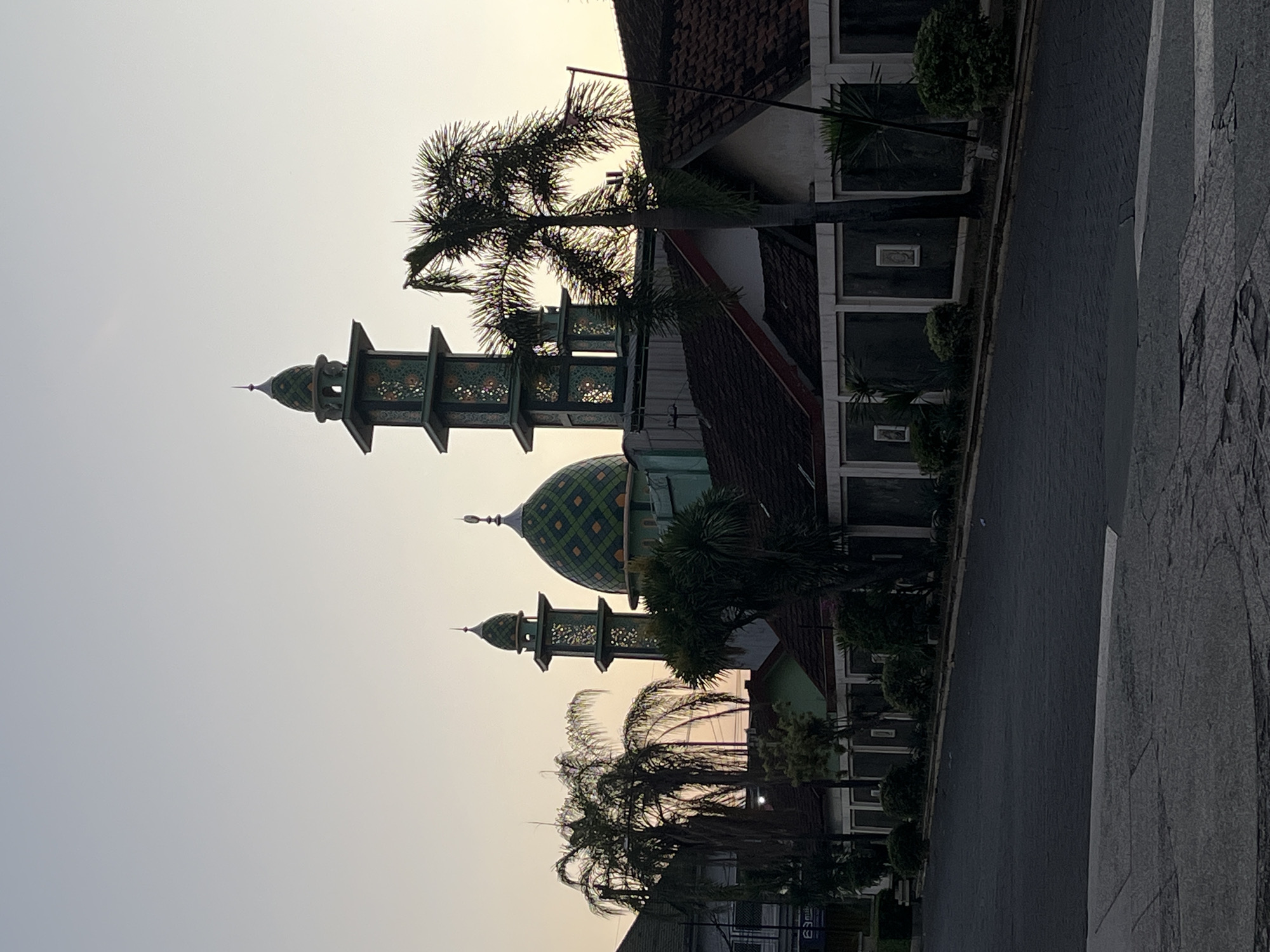 In Boro, Indonesia, a mosque with green domes and minarets stands against a pale, hazy sky, possibly at dawn or dusk. The mosque is flanked by palm trees and other greenery. In the foreground, a dark asphalt road leads away from the viewer, with a tiled pavement on the right side. The scene appears to be in a residential area, with houses and a wall lining the street. There are no people or animals visible in the image.