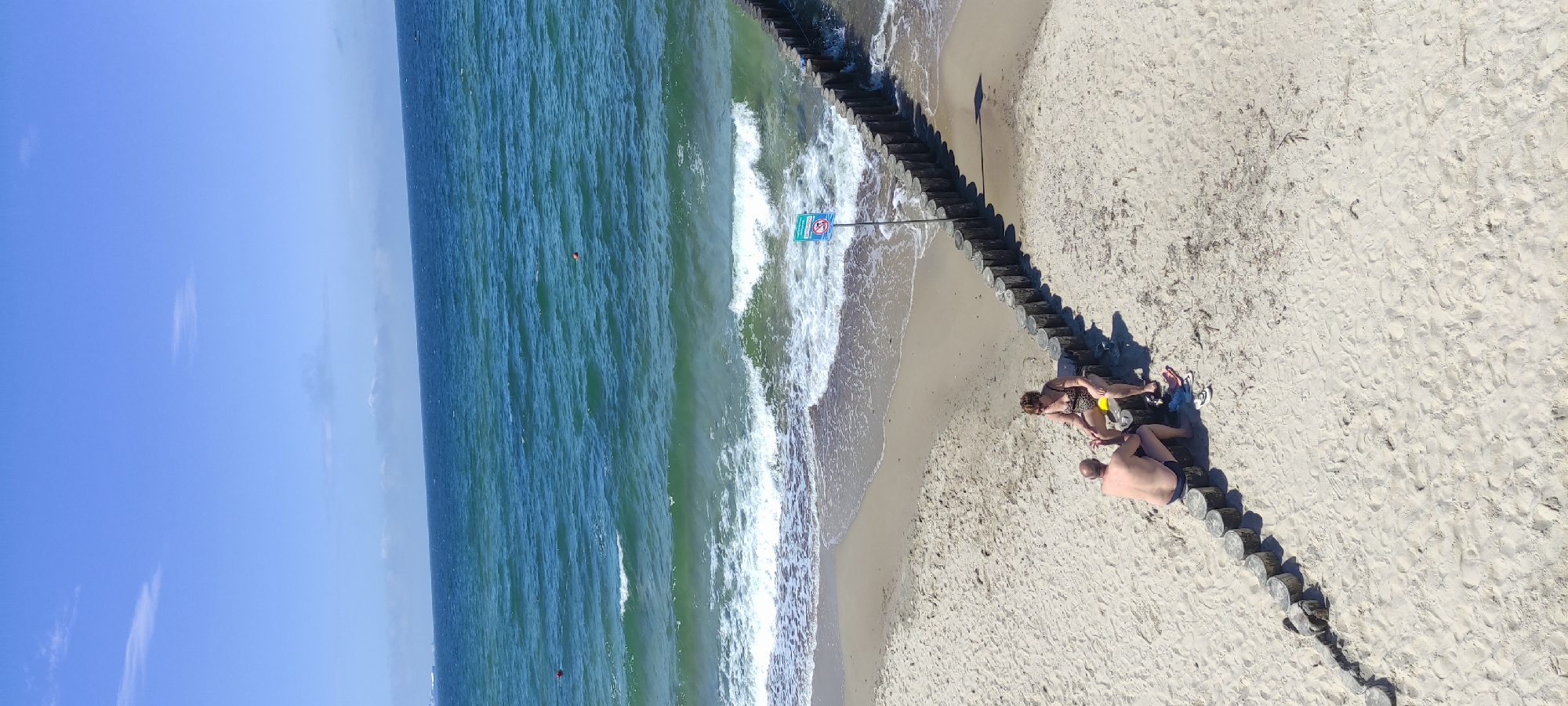 Two individuals, a male and a female, are seated on a wooden structure on a sandy beach in Zelenogradsk, Russia. The male, wearing dark swim trunks, is facing away from the camera. The female, wearing a patterned bikini and sunglasses, is seated next to him and appears to be looking at him. They are positioned on a breakwater made of rounded wooden logs that extends into the sea. The sea, with moderately choppy, green-tinted water and breaking waves, occupies the upper portion of the frame. The sky above is clear blue with some distant, wispy clouds. A sign with a red circle and a diagonal line through a silhouette is visible near the breakwater. Several buoys are visible in the water. The beach is sparsely populated with scattered debris on the sand.