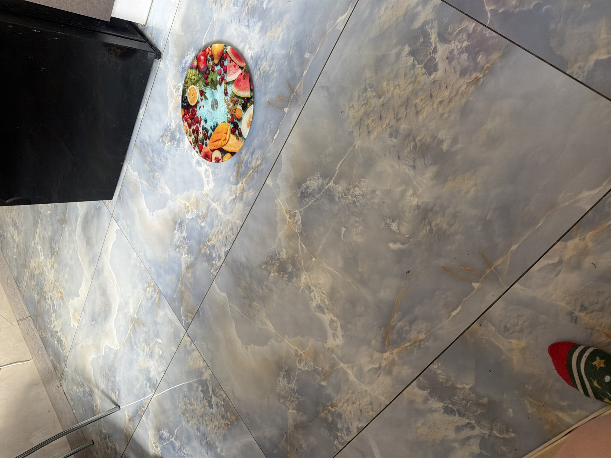 The image shows a floor with large, marbled tiles in shades of blue, gray, and beige. In the upper right portion of the image, a portion of a black appliance, possibly a refrigerator, is visible. In the center of the floor, a round mat with a colorful fruit pattern is placed. The mat features various fruits such as strawberries, blueberries, grapes, oranges, mangoes, watermelon, and peaches arranged on a light blue background. In the bottom left corner of the image, a person's foot wearing a sock with a festive pattern of stars and stripes is visible, with the toe pointing towards the fruit mat. The scene appears to be indoors, likely a kitchen or dining area. The location is not specified in the image.