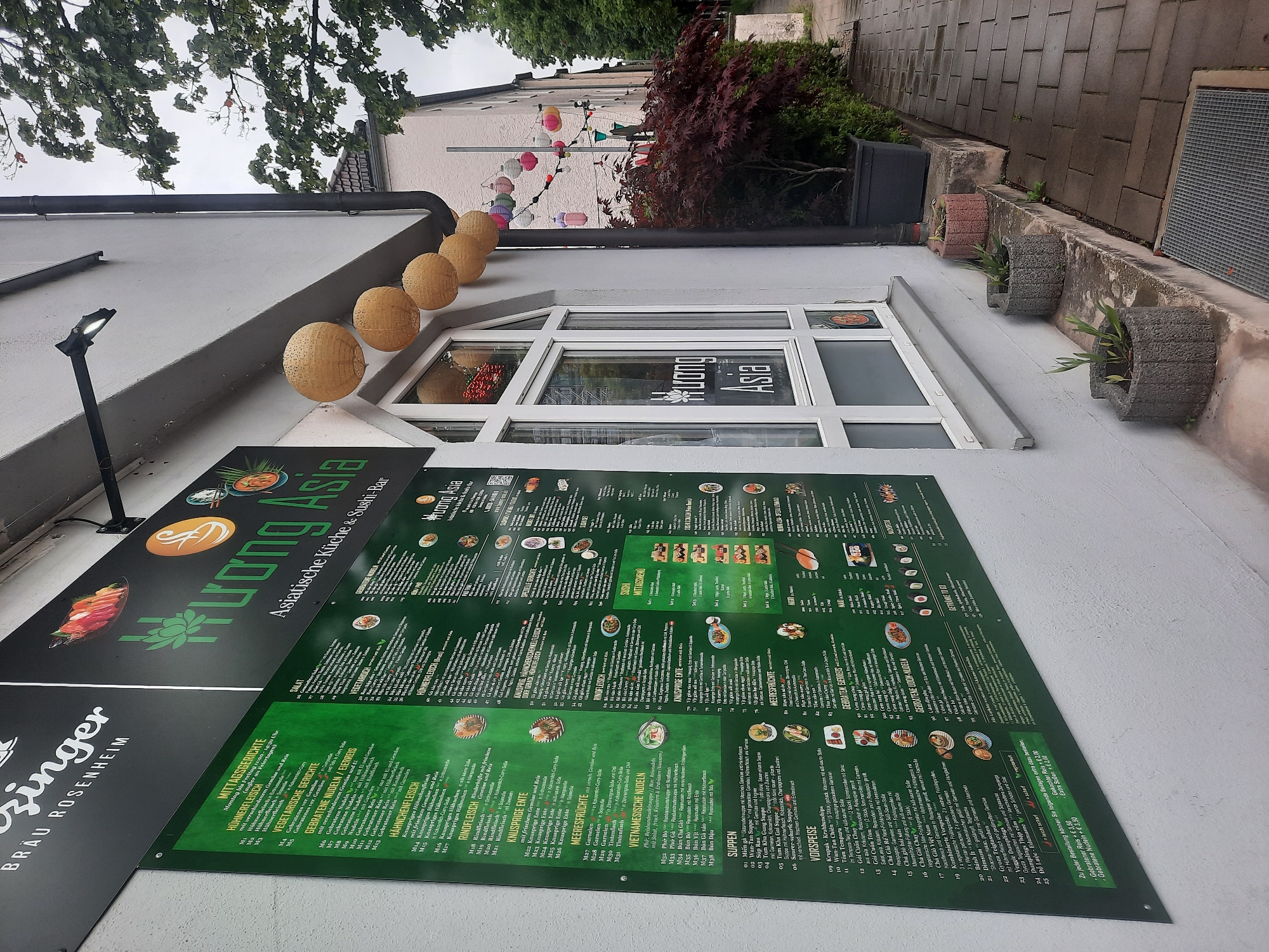 The image shows the exterior of an Asian restaurant named "Huong Asia" in Rosenheim, Germany. The restaurant has a large green menu board mounted on the wall, displaying various dishes and prices. The menu is in German and lists categories such as "Mittagsgerichte," "Vegetarische Gerichte," "Gebratene Nudeln / Eiernudeln," "Hähnchenfleisch," "Rindfleisch," "Knusprige Ente," "Meeresfrüchte," and "Vietnamesische Nudeln." There are also sections for "Suppen," "Vorspeise," and "Sushi."
The restaurant's name, "Huong Asia," is prominently displayed in a stylized font on the menu board and also on a sign above the window. Paper lanterns, some in white and others colorful, are strung across the facade and hanging from the building. A large window with a white frame provides a view into the restaurant.
The building appears to be a typical German building with a light-colored facade. In front of the restaurant, there is a sidewalk paved with grey rectangular stones. Several planters containing plants are arranged along the wall. A dark-colored drainpipe runs down the side of the building. Trees with green foliage and a tree with dark red leaves are visible in the background. The sky is overcast, suggesting a cloudy day. The image is taken from a slightly low angle, looking up at the restaurant and its signage.