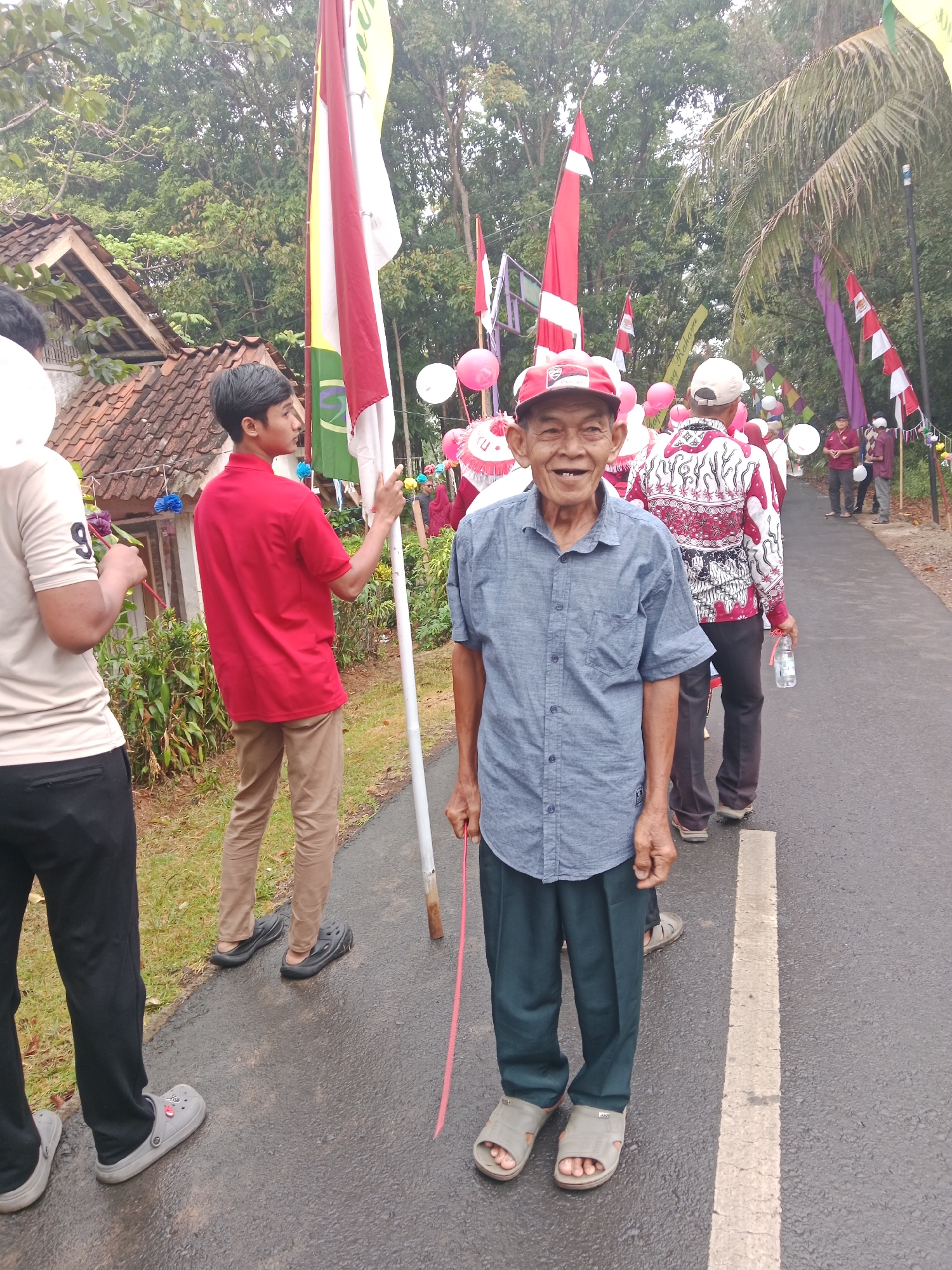 In Bojongmengger, Indonesia, a group of people participate in a parade on a wet asphalt road. The foreground is dominated by an elderly man smiling at the camera, wearing a blue collared shirt and dark pants. He holds a red stick. Behind him, a young man in a red polo shirt and khaki pants carries a flag with red and white stripes. Other participants, some in traditional Indonesian clothing, are visible in the background, holding flags and balloons. The environment includes lush green trees and vegetation, a tiled-roof building, and Indonesian flags displayed along the road.