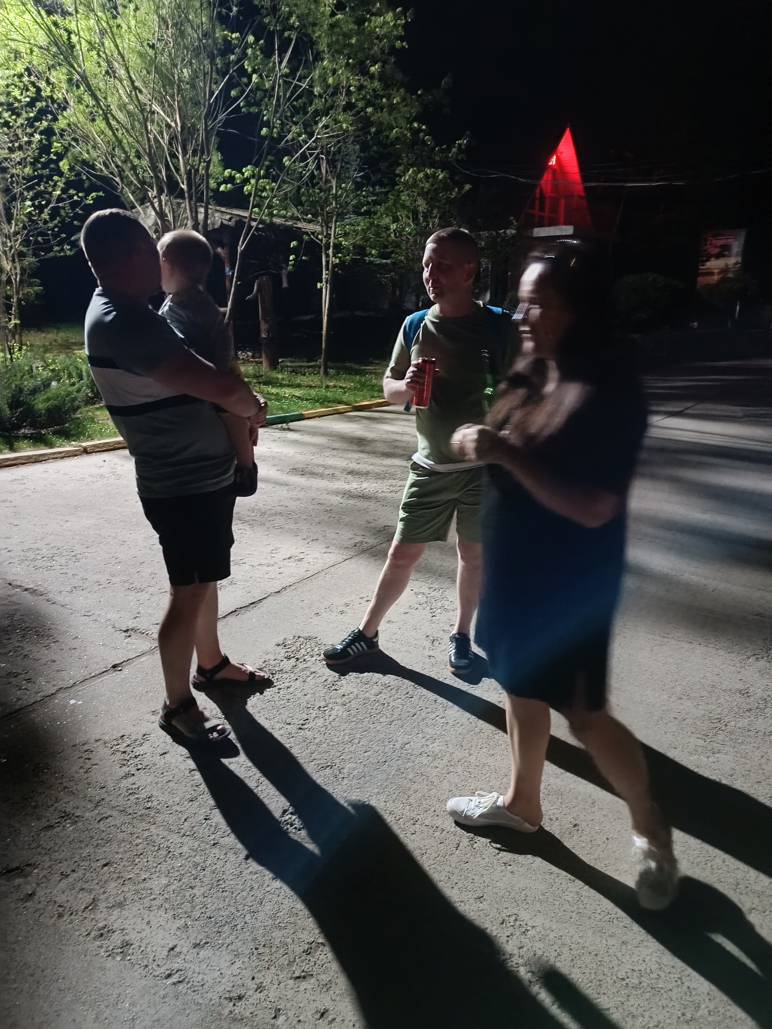 A group of four people is gathered outside on a paved area, likely a street or courtyard, at night. A man is holding a toddler. Beside them, a second man stands with a red can in his hand. A fourth person, an adult female, is walking past the group.
In the background, there are trees and some structure with red triangular lights.
The location is likely in Chaykovsky, Russia.