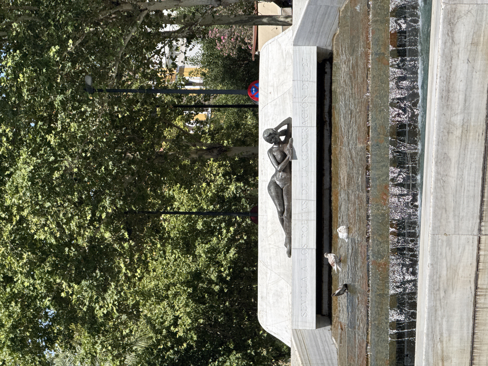 The image shows a sculpture of a woman lying down on a marble pedestal, with water flowing down steps in front of it. The sculpture appears to be made of bronze and depicts a nude female figure in a relaxed pose, with her head resting on her hand. The marble pedestal has text carved into it, which reads "SEVILLA A LOS POETAS DE LA GENERACION DEL 27". This translates to "Seville to the poets of the Generation of '27", indicating the sculpture is a tribute to poets from that era.
The scene is outdoors, with lush green trees and foliage visible in the background. Sunlight filters through the leaves, suggesting it is daytime and the weather is clear. Two pigeons are perched on the edge of the water feature, and a third pigeon is standing near them. The water is clear and appears to be flowing gently down the stone steps. The overall impression is one of a tranquil and artistic public space, likely a park or plaza in Seville, Spain. The bright sunlight and vibrant greenery suggest a pleasant, warm day.