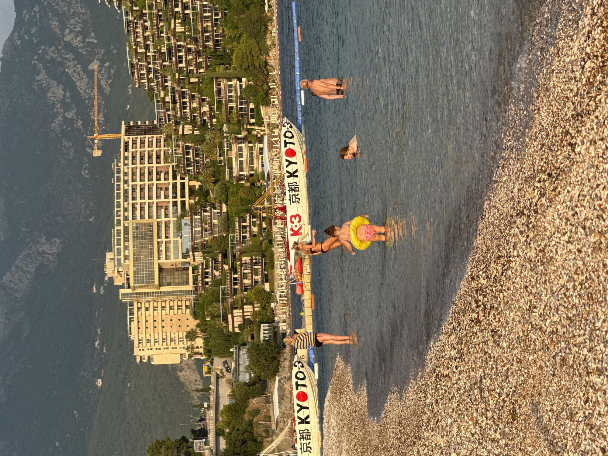 This image depicts a sunny day at a pebble beach in Budva, Montenegro. The scene is dominated by the calm, blue sea and a densely built hillside with modern apartment buildings adorned with greenery. The time of day appears to be late afternoon, judging by the warm, golden light casting shadows and illuminating the pebbles on the shore.
Several people are enjoying the water. In the foreground, a person wearing a striped shirt and black shorts stands in the shallow water, looking towards the left. Further out, two women are visible: one wearing a yellow inflatable ring is lying on her back, while another is sitting in the water. A man is also present in the water, partially submerged, and appears to be swimming.
A boat with prominent Japanese text is beached on the shore. The text includes "京都" (Kyoto) and "KYOTO" in English, along with the character "3". A yellow construction crane is visible on the land behind the buildings, suggesting ongoing development or construction in the area. The overall atmosphere is one of leisure and relaxation, with the warm sunlight and gentle water contributing to a peaceful ambiance.