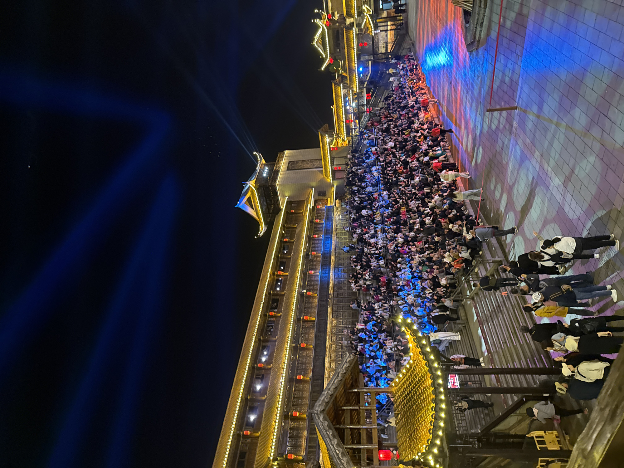 This media file captures an evening scene in Wulingyuan, China, depicting a large outdoor gathering attending a performance or show. The image is taken at night, with the dark sky illuminated by numerous artificial lights.
The main subjects are a dense crowd of people, most of whom are seated in an open area, facing towards a performance space. In the foreground, additional people are standing, also observing the event. The crowd extends across a significant portion of the scene, indicating a popular public event.
The setting is characterized by elaborate, multi-story buildings constructed in a traditional Chinese architectural style. These structures are richly adorned with golden decorative elements, intricate roofs, and numerous red lanterns, which glow warmly against the dark backdrop. Beams of blue light emanate from sources behind the buildings on the left, cutting across the night sky. On the ground in the performance area, vibrant blue, red, and orange lights are projected, creating a dynamic and colorful display. High above the buildings on the left, an aerial performer or prop, possibly suspended by wires, is visible, adding to the spectacle.
The activity taking place is clearly an entertainment event, likely an outdoor stage show or cultural performance, given the large, attentive audience, the theatrical lighting, and the aerial elements. The atmosphere appears lively and engaging.
Notable details include the clear night sky, the extensive and decorative illumination of the traditional Chinese buildings, and the intense, colorful lighting used for the performance. The architectural style, combined with the location context provided, confirms the Chinese setting.
Visible text includes some Chinese characters on vertical red banners decorating the buildings, though the resolution makes specific transcription difficult.