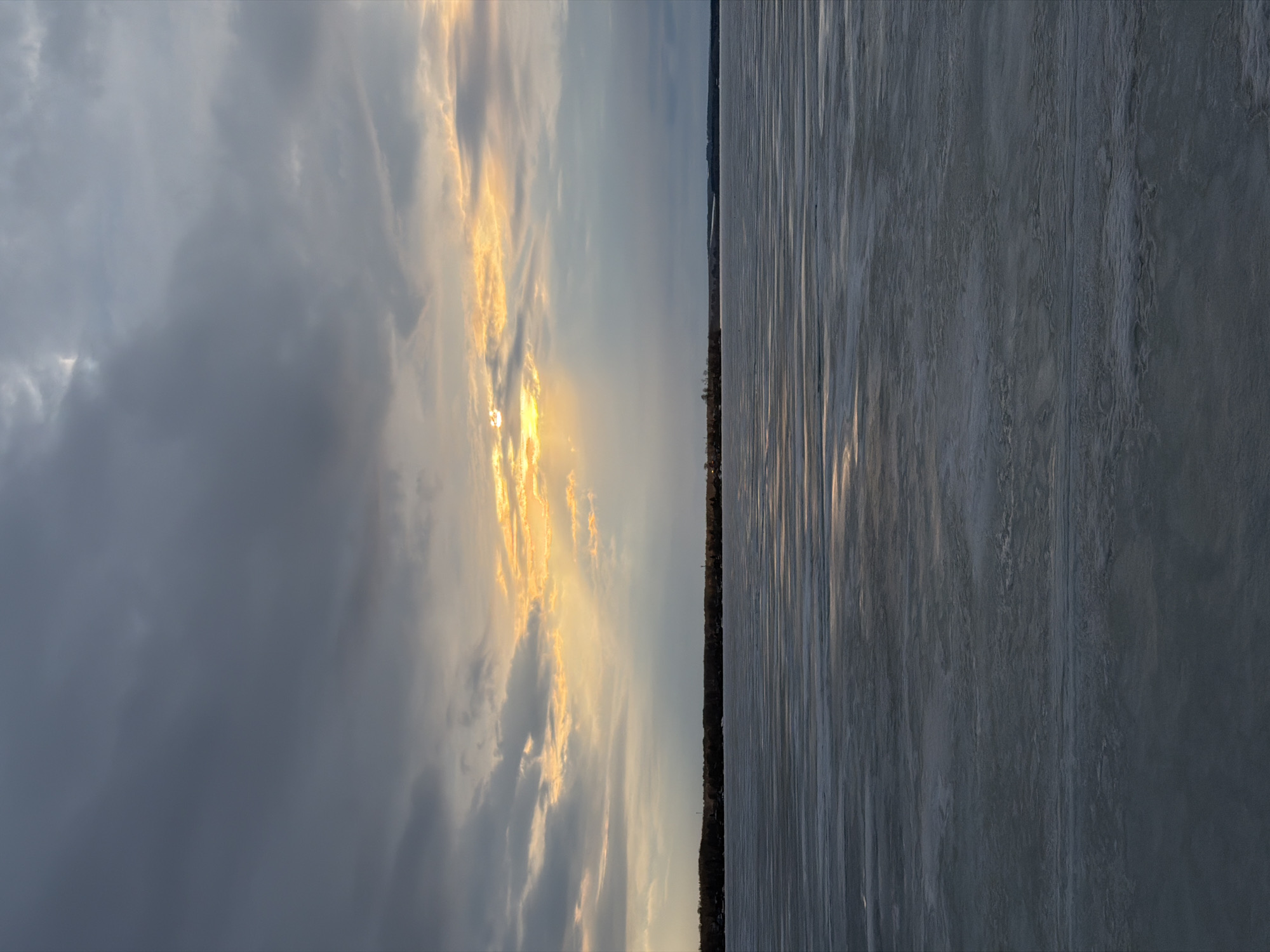 The image depicts a landscape scene, likely taken in Votkinsk, Russia, at or around sunset. The left side of the image features a sky filled with dense, gray clouds, transitioning to lighter, sunlit clouds with golden hues closer to the horizon. A hint of the sun is visible through a gap in the clouds, suggesting a sunset or sunrise. The right side of the image shows what appears to be a frozen body of water, potentially a lake or reservoir. The ice surface has a rough texture with visible striations and reflections of the sky. A dark line of trees or buildings is visible on the horizon, separating the sky and frozen lake. There are no people visible in the image. The environment suggests a cold, possibly winter, setting.