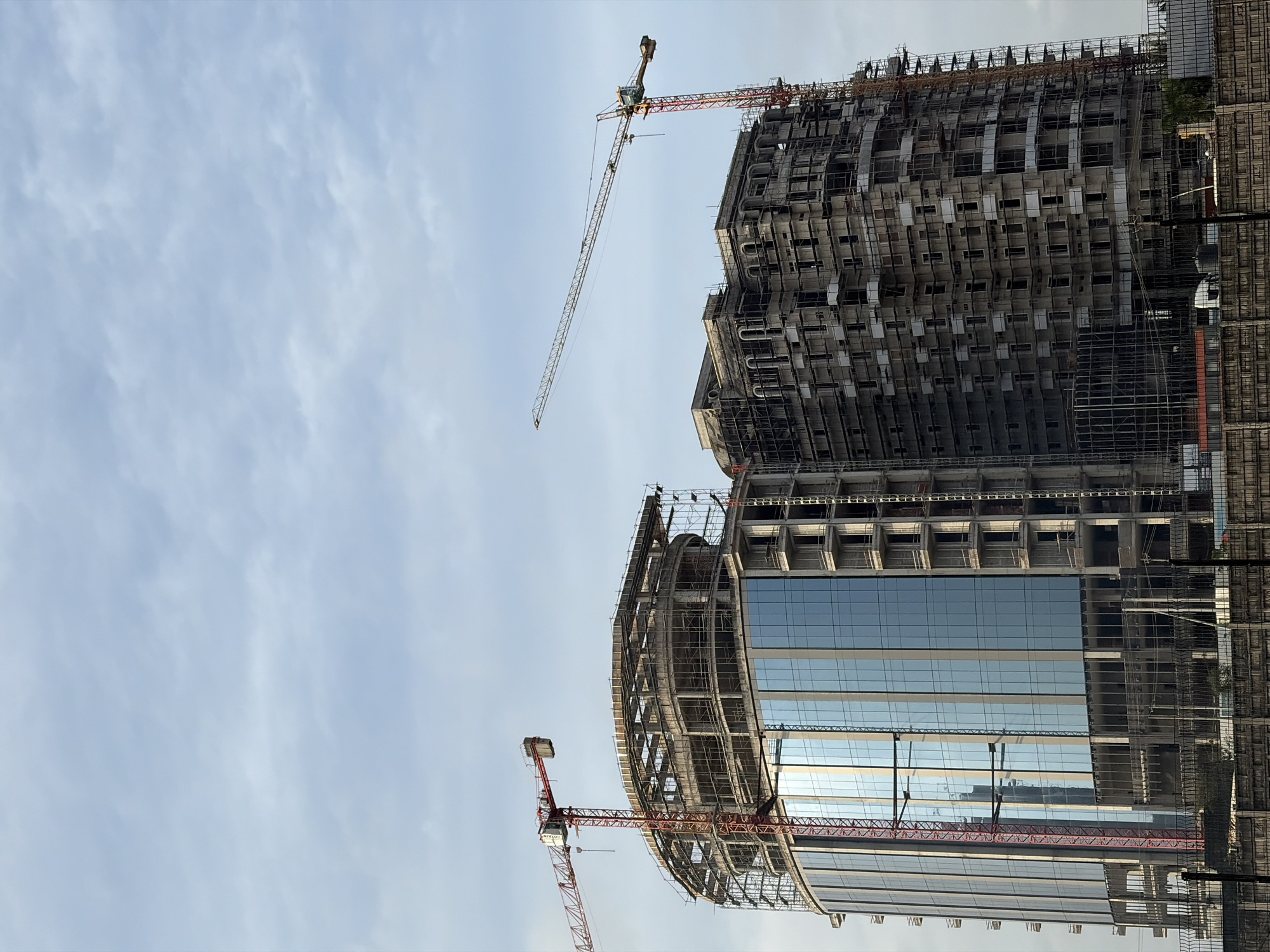 This is a full-frame, eye-level, outdoor, daytime photograph of two high-rise buildings under construction in Alaguntan, Nigeria. The sky is overcast with clouds. The building on the left is a rounded structure with blue glass windows and exposed concrete framework on its upper floors, where scaffolding is visible. A red tower crane is positioned next to it, extending over the building. The building on the right is a rectangular structure covered in scaffolding, with visible concrete floors and arched window openings on its upper facade. A yellow and red tower crane is situated behind this building, with its boom extending horizontally. In the foreground, a dark metal fence runs across the bottom of the frame, behind which are some dark structures and what appears to be a white vehicle.