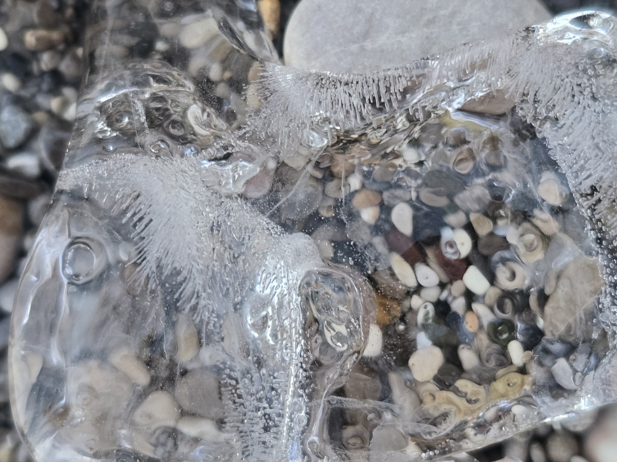 The image shows a close-up, macro view of ice formed around pebbles on a beach. The ice is transparent with many trapped air bubbles and crystalline formations, creating intricate patterns. The pebbles beneath are visible through the ice and are of various sizes, shapes, and colors, including gray, white, brown, and some with darker markings. The background is blurred but appears to be a continuation of the pebble beach. The lighting is bright, highlighting the textures and details of the ice and pebbles. The image does not contain any people, actions, or interactions. The location in the city of Salerno, Italy, is not discernible from the visual information presented in the image.