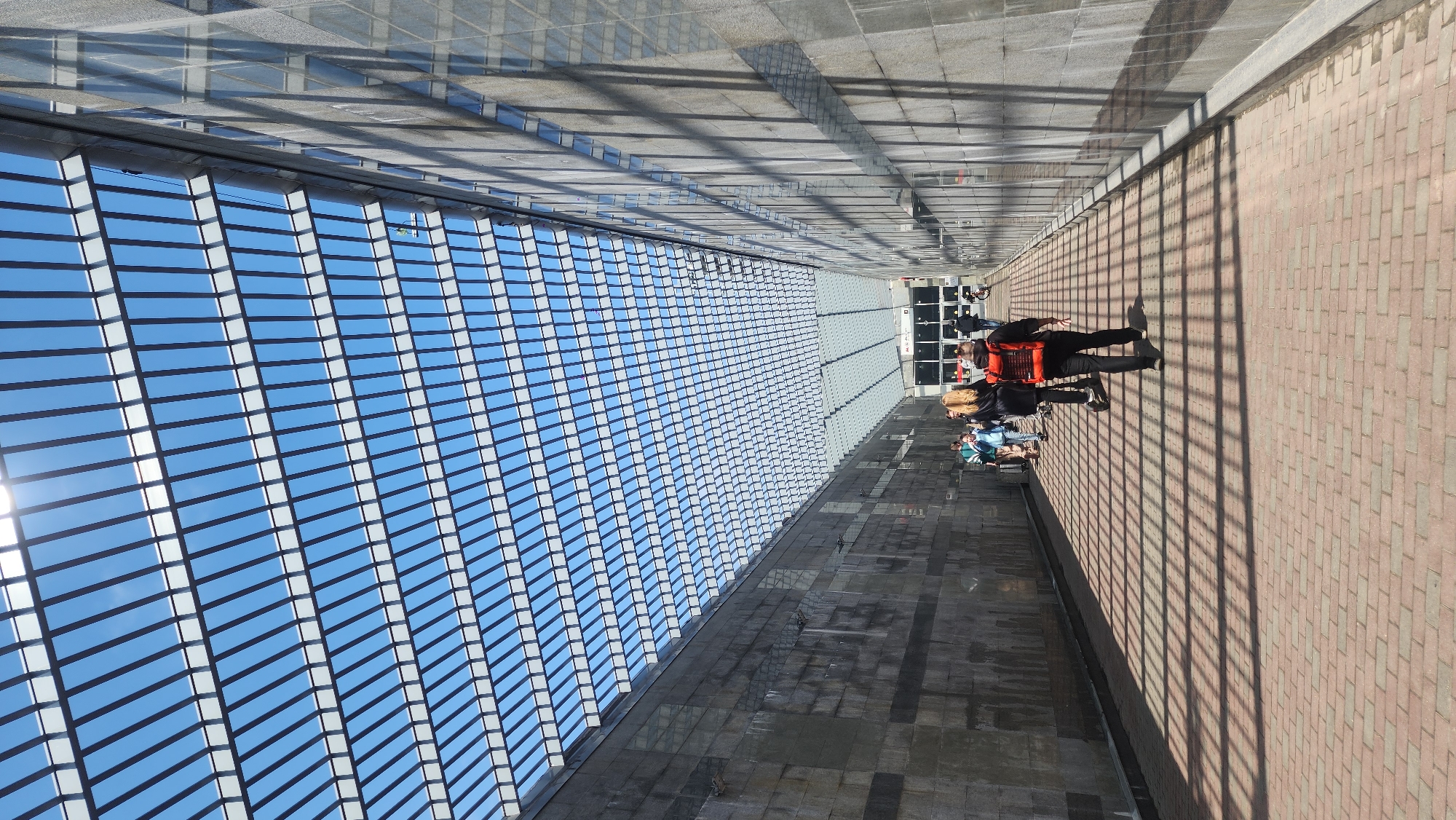 A long, horizontal outdoor shot features a partially-covered walkway next to a brick road and wall in an urban setting. The covered walkway has a lattice roof creating a grid of long rectangular beams that allow a view of the sky between each space. These beams cast shadows on the walkway. The walkway is gray tile, next to a brick road on the right. On the road, shadows from the overhead structure cast linear lines across the surface. A group of people are clustered together on the walkway, near the end of the covered area. The people are a mix of ages and genders, and some are wearing jackets.