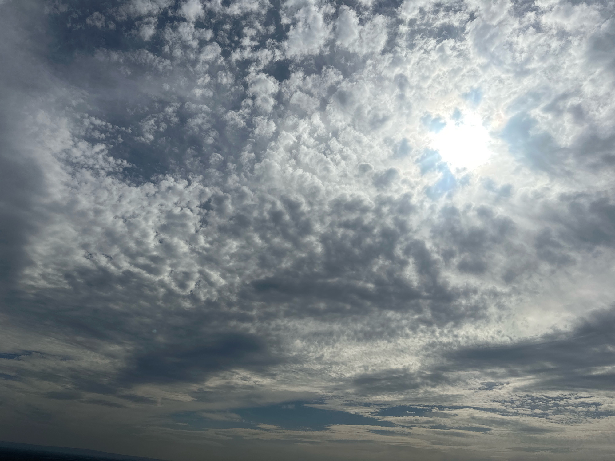 The image depicts a sky filled with a variety of clouds. The sun is visible through a gap in the clouds in the upper right quadrant, casting bright rays. The clouds are a mix of white and gray, with some appearing wispy and others more dense. The lower portion of the image shows a hazy, indistinct landscape that could be a city or rural area, with a sliver of dark land visible along the horizon. There are no people, objects, or actions depicted in the image. The location is identified as Simskii, Russia.