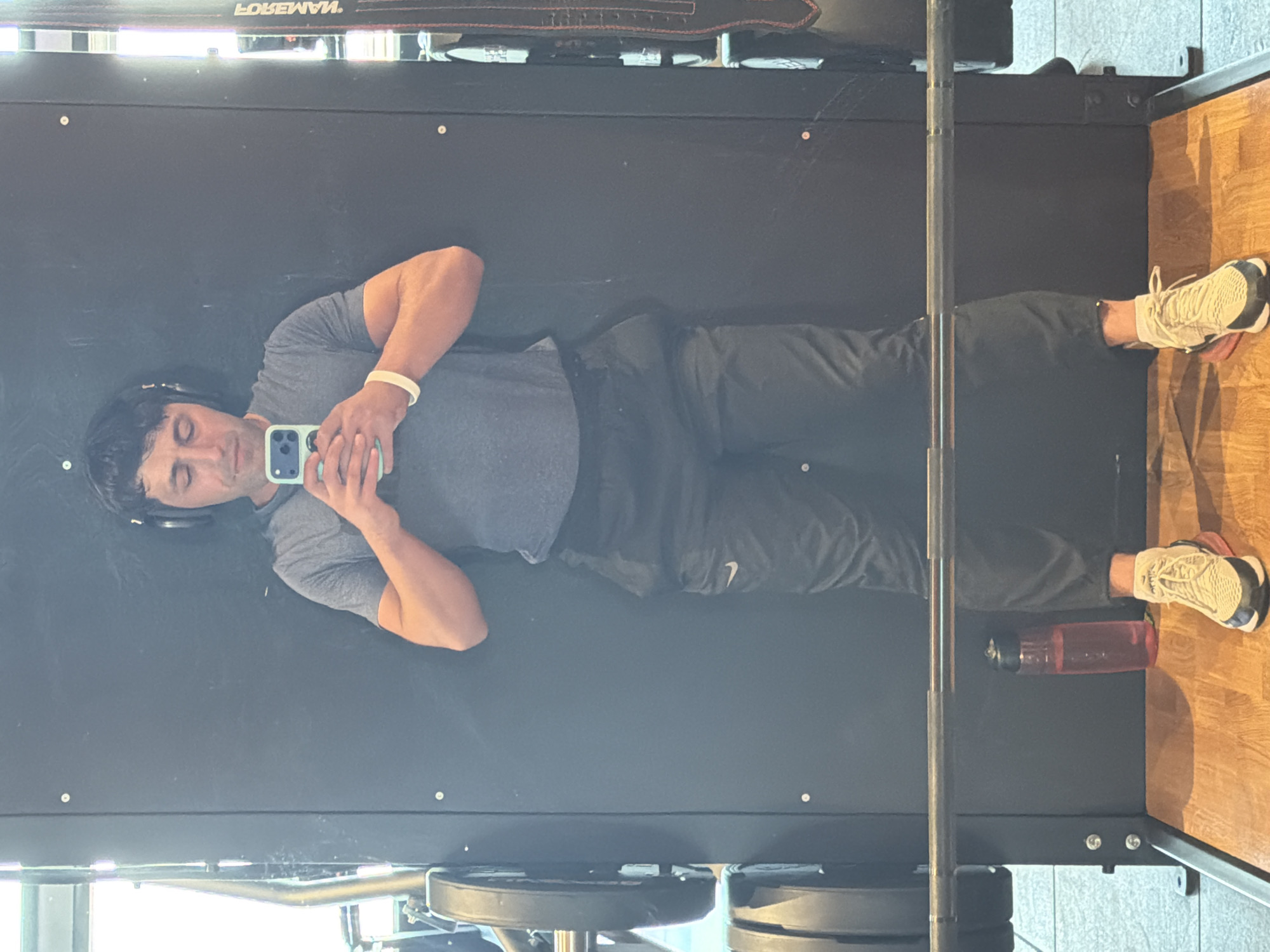 The image is a mirror selfie taken by a young man lying on gym equipment in what appears to be a fitness center in Krasnoyarsk, Russia. The man, seen from the waist up, wears a grey short-sleeved t-shirt, black athletic pants with a white Nike swoosh on the left thigh, a white wristband on his right wrist, and black over-ear headphones. He holds a smartphone with a light blue case in both hands, angled to capture his reflection. His expression is neutral and relaxed, suggesting he might be taking a break from a workout.
He is lying on a dark, padded surface, which is part of a larger gym machine. A red water bottle rests next to him on this surface. The floor visible near his feet transitions from the dark equipment surface to a wooden flooring section. Heavy, circular weight plates are partially visible in the bottom portion of the frame, further indicating the gym environment. In the background, some reversed text, possibly "WAVEMVIOA" or a similar brand name, is discernible as a reflection on the equipment or a wall. The scene is brightly lit, consistent with an indoor daytime setting.