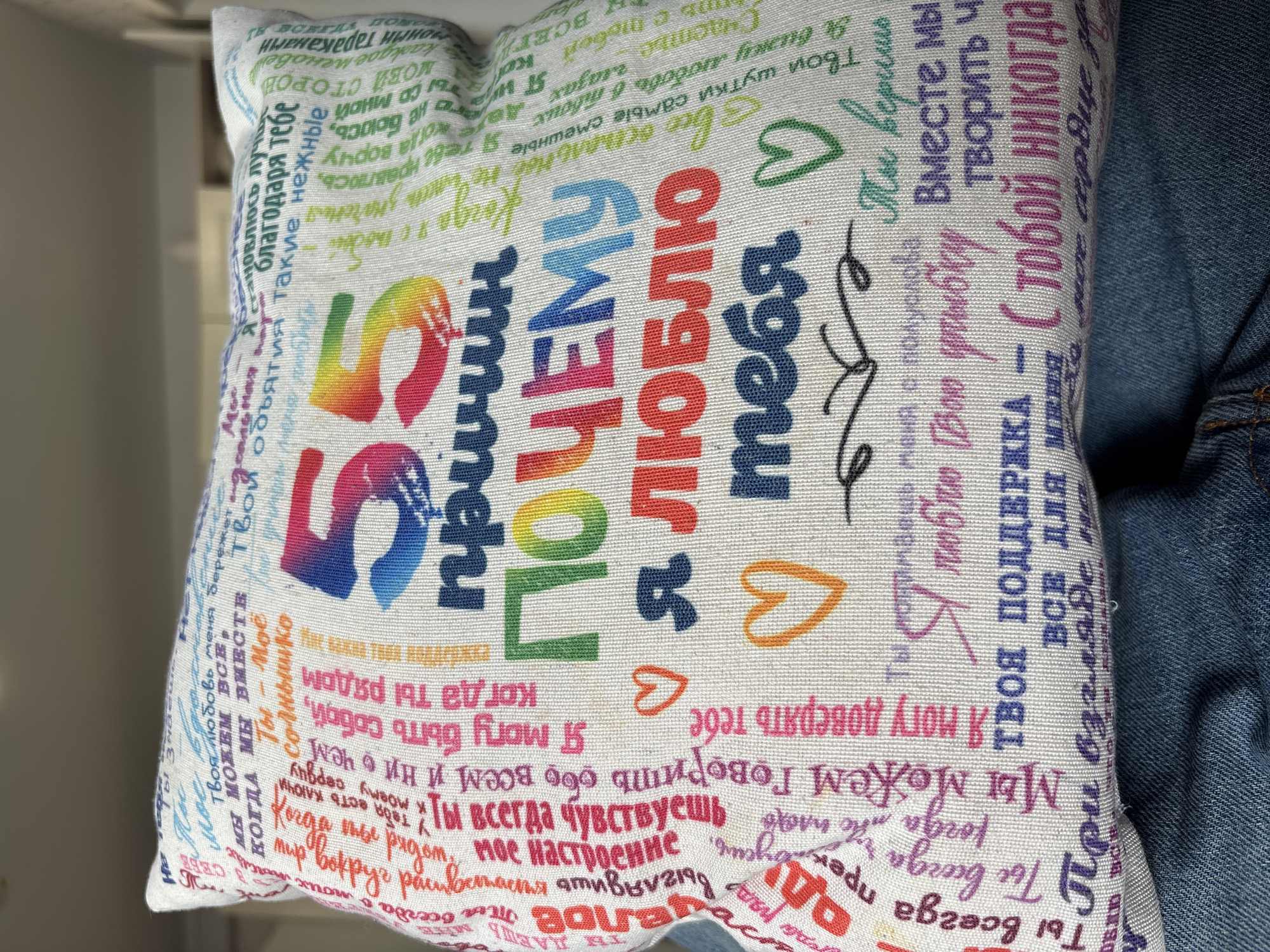 A close-up shot shows a decorative pillow placed on top of a pair of blue jeans. The pillow is white and covered in colorful text and heart illustrations in various fonts and sizes. The text appears to be in Russian, expressing sentiments of love and appreciation. The letters "55 причин" (55 reasons) are prominently displayed in rainbow colors. The background is blurred, but a wall and part of a window frame are visible. The jeans are lightly textured with visible seams and stitching. The overall impression is a casual, indoor setting. There is no explicit reference to Izhevsk, Russia, in the image.