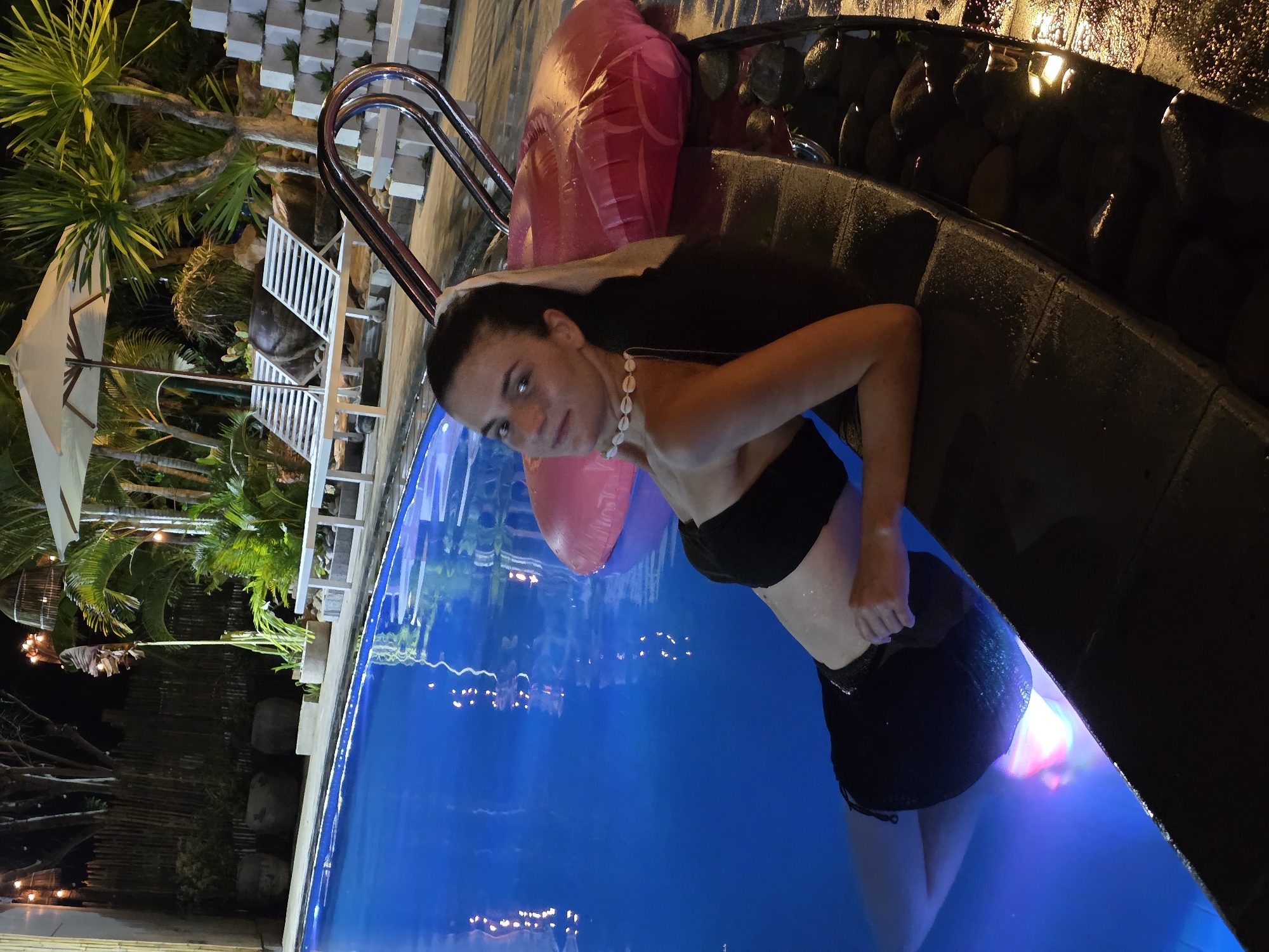 A woman wearing a black bikini top and black shorts is standing in a swimming pool at night in Mũi Né, Vietnam. She has dark hair and is wearing a white seashell necklace. A large pink inflatable ring is floating behind her. The pool is lit with blue and white lights. There is a pool ladder and lounge chairs to the left of the pool. Tropical plants are visible behind the lounge chairs. The edge of the pool is lined with dark stones.