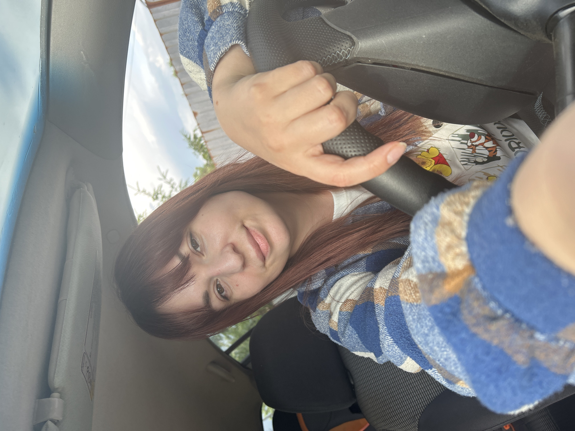 A woman is in a car, holding the steering wheel. She is wearing a blue and tan plaid shirt over a white top with Winnie the Pooh characters. The inside of the car is visible, including the dashboard, seat, and sun visor. Outside the car, there are trees and a building under a cloudy sky. The location cannot be confirmed as the city of Фоки, Russia.