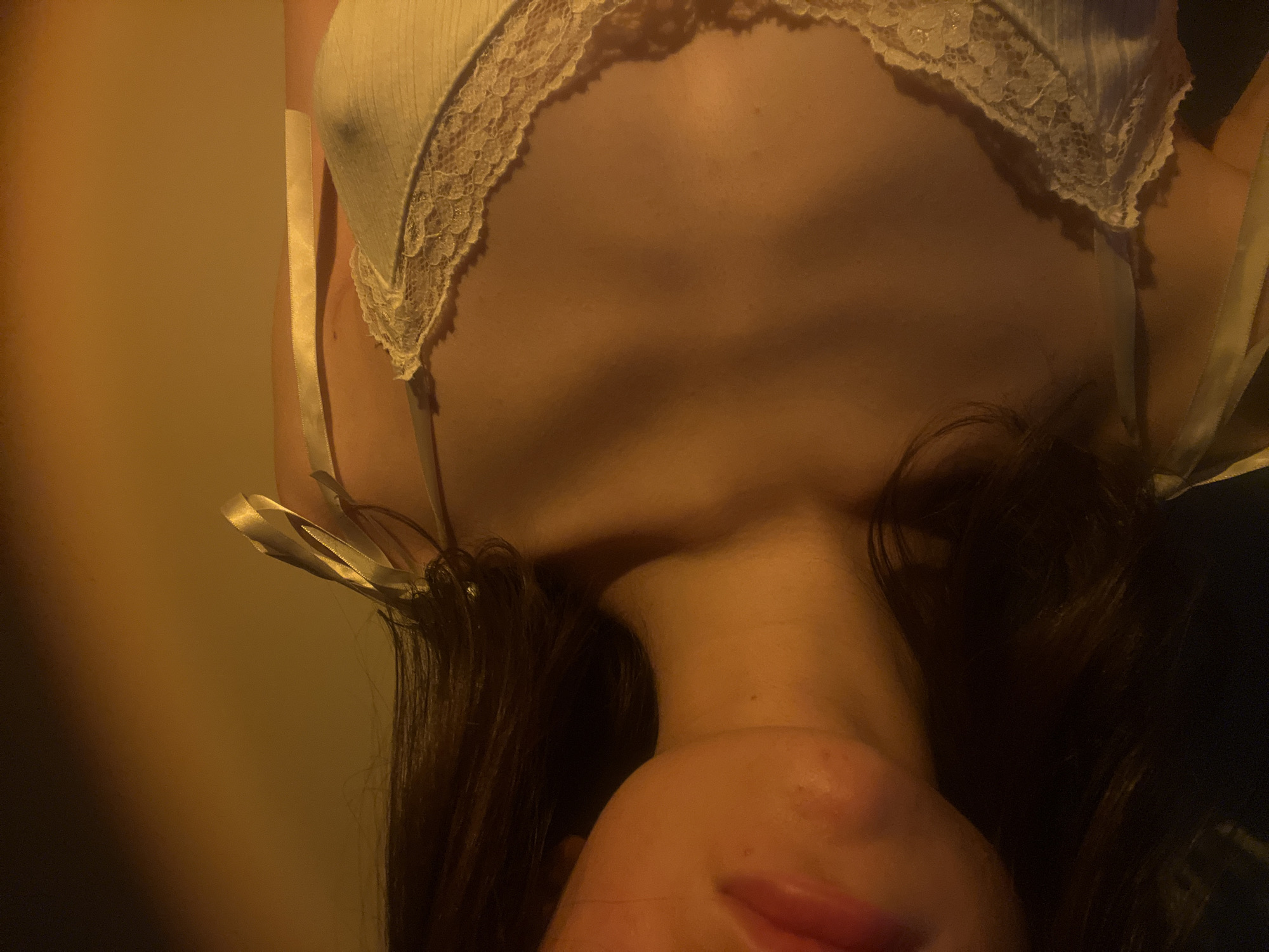 The image shows a close-up of a person's upper chest and neck area, with their face partially visible. The person is wearing a light-colored top with lace detailing along the neckline and ribbon straps. The lighting is warm and atmospheric, suggesting a low-light environment. There is no indication of the location within the city of Ternopil, Ukraine, or any specific objects or actions that would place it in that city. The image is a portrait focusing on the texture and details of the clothing and skin.