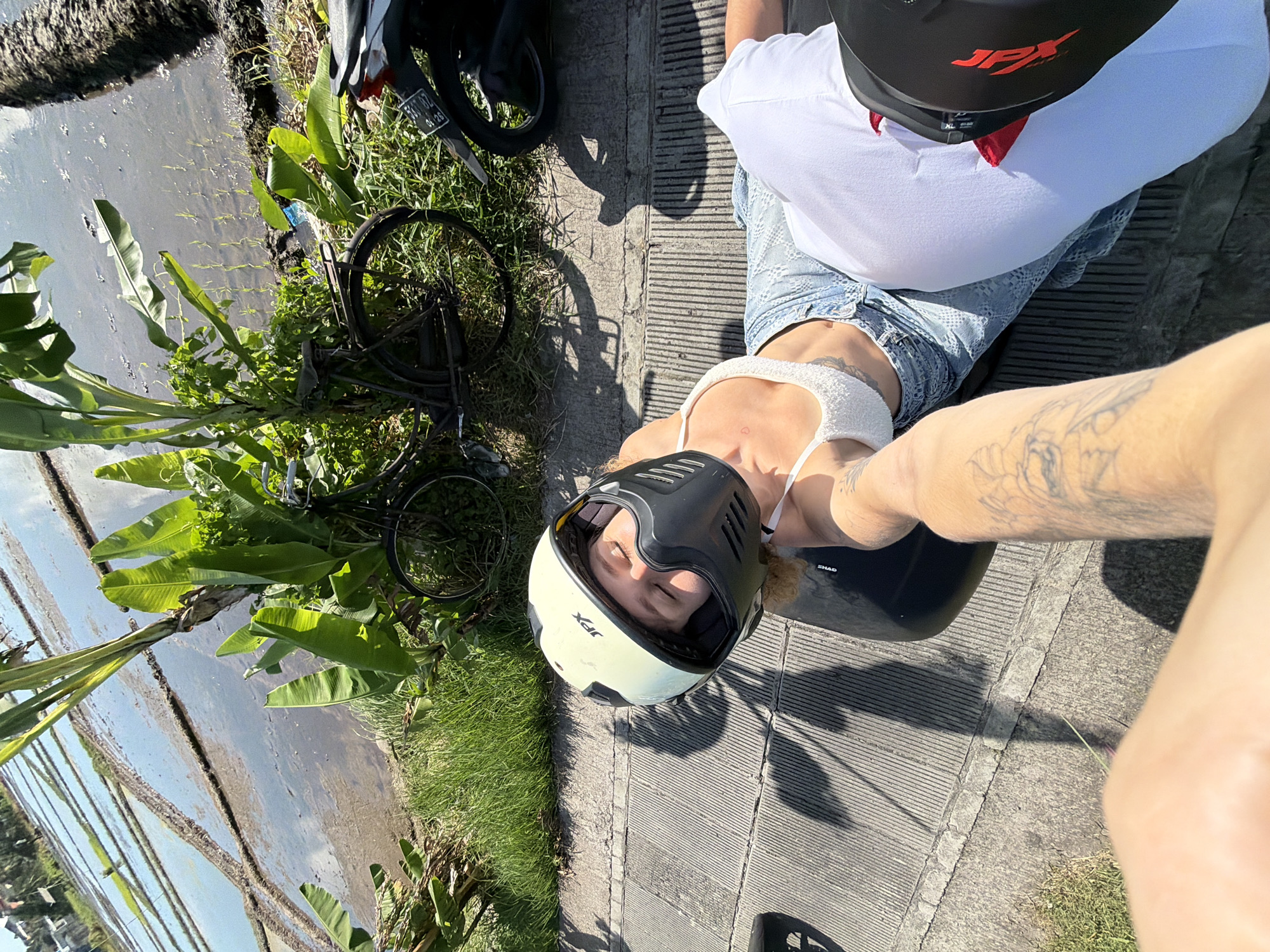 A high-angle perspective captures two individuals, one seemingly taking a selfie. A woman wears a white crop top and distressed denim shorts. She has a white and black helmet on. Another person is visible behind her in a white T-shirt, and they're also wearing a black helmet. They are positioned on a gray concrete pathway.
Adjacent to the pathway is a grassy area with a bicycle resting against it. Beyond the grass, a rice paddy terrace stretches into the distance, reflecting the sky. A motorcycle is partially visible near the bicycle. The scene appears to be in North Kuta, Indonesia.