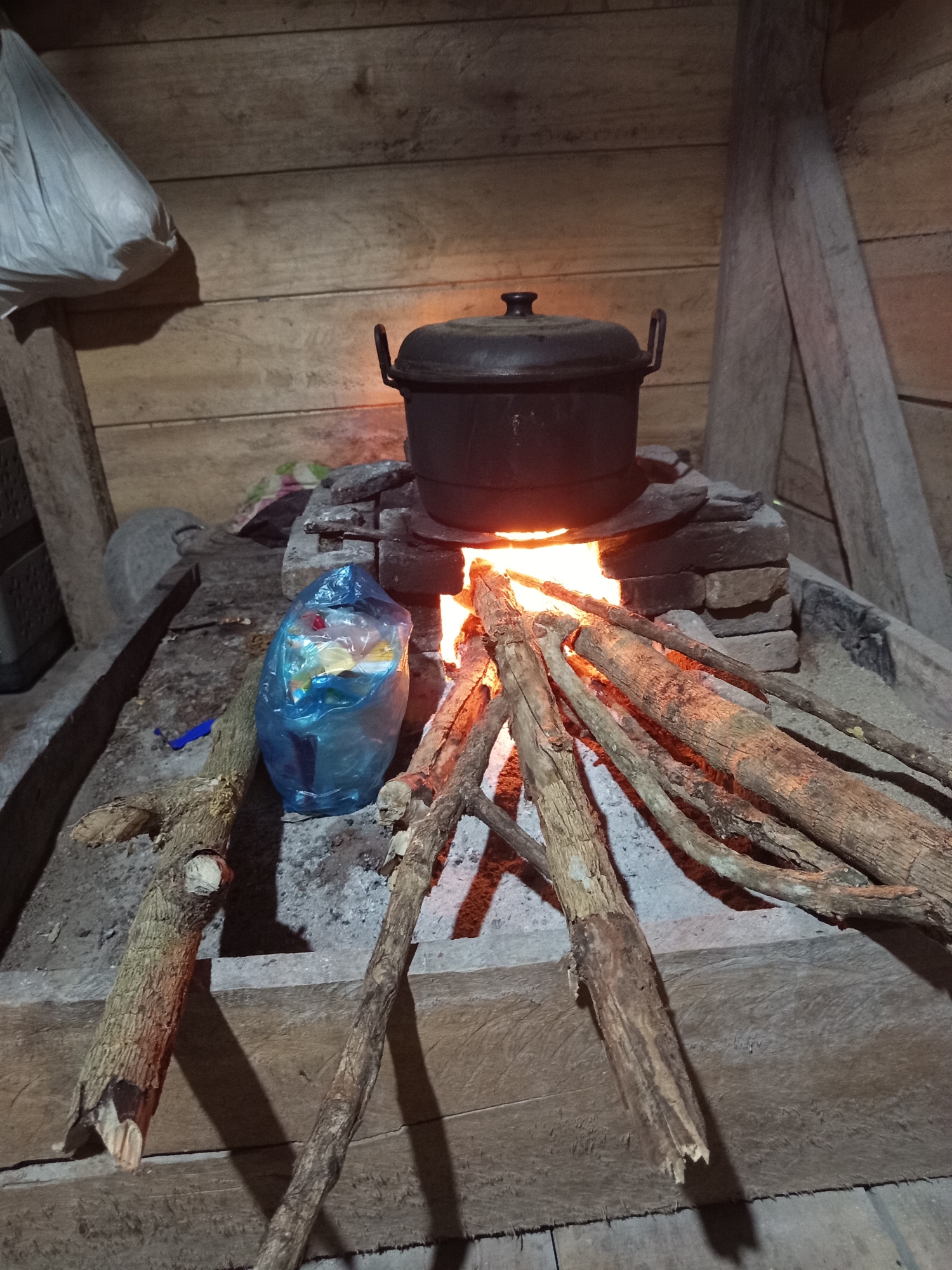 An outdoor scene in an Indonesian village shows a rustic cooking setup. A black pot with a lid sits atop a makeshift stove constructed from stones and bricks, with a fire burning underneath, emitting a warm orange glow. Several pieces of firewood are arranged around the fire. To the left of the stove, a blue plastic bag contains various items. The background is made of wooden planks, suggesting a simple dwelling or structure. The ground is covered in a sandy or ashy material, with more logs scattered around. The overall impression is one of a humble and traditional cooking environment.