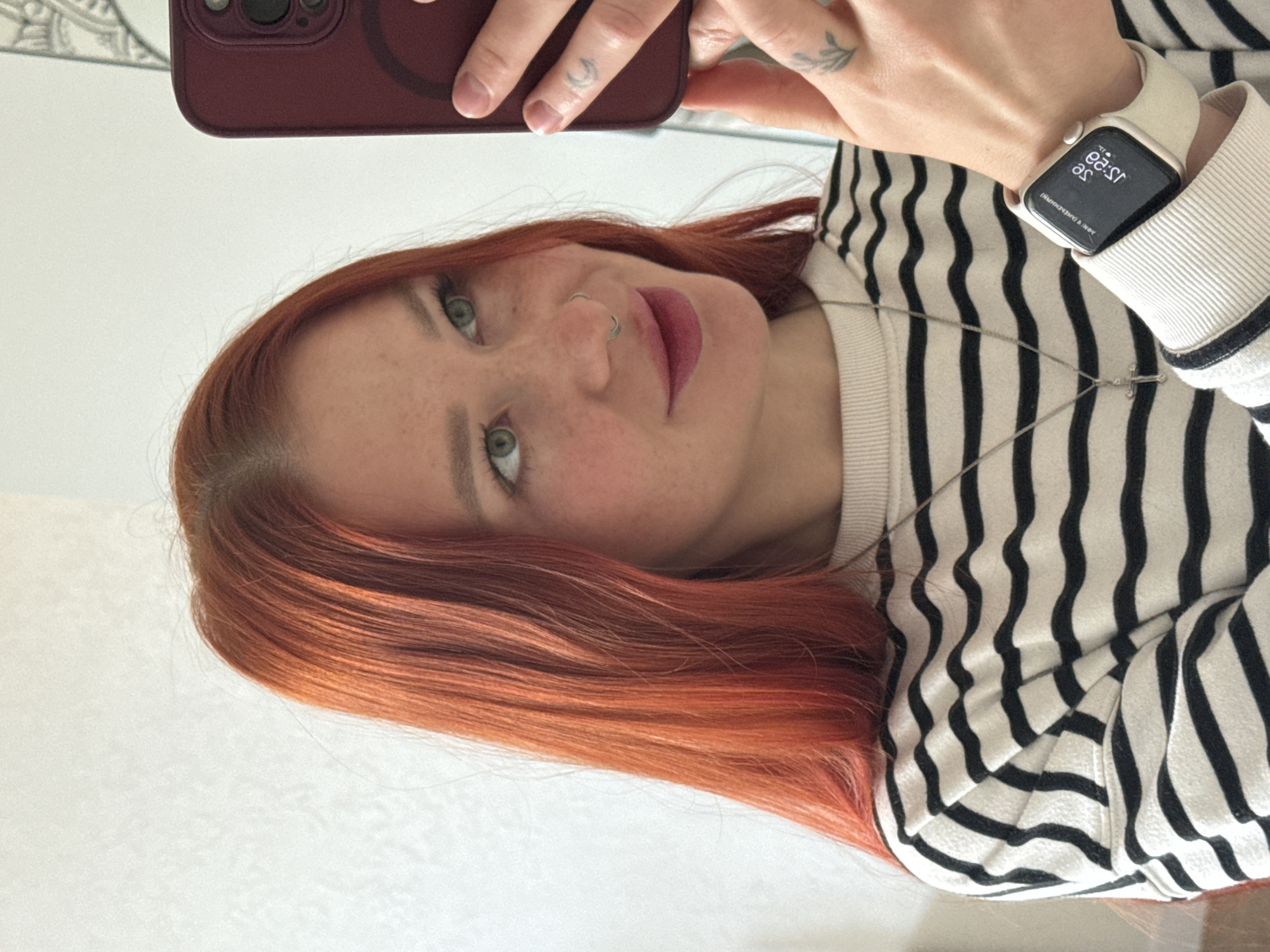 A fair-skinned woman with red hair and freckles takes a selfie with a phone in a burgundy case. She wears a black and white striped sweater and a silver necklace with a cross. On her left wrist is a white smartwatch. She has a nose ring and tattoos on her fingers and wrist. The scene does not provide information about the location in the city of Rostov-on-Don, Russia.