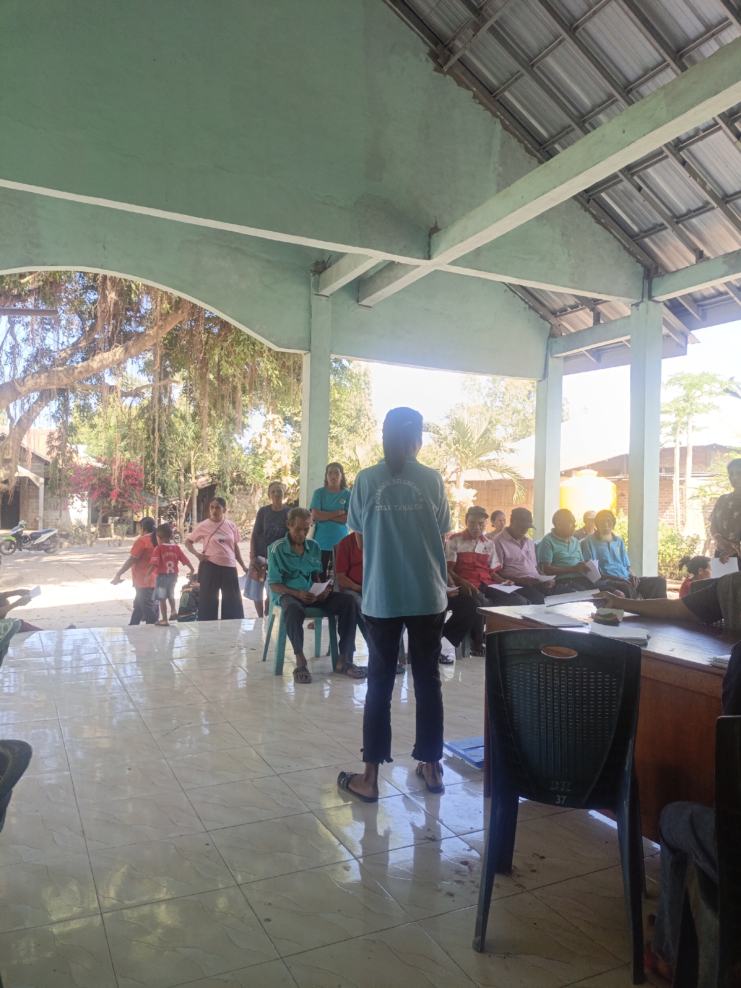 The image depicts an outdoor gathering at what appears to be a community hall or open-air pavilion in Tanah Lein, Indonesia. The structure has a light green painted ceiling and supporting pillars. Sunlight streams in from the right, illuminating the tiled floor. A group of approximately 10-15 individuals, including adults and children, are present. Some are seated on plastic chairs, while others are standing. One person, wearing a blue t-shirt with text that reads "POSYANDU KELUARGA BUNDA DESA TANALEIN," stands facing away from the camera, appearing to address the seated individuals. Several people are holding papers. In the background, there are trees with visible roots, a motorcycle, and buildings. A yellow water tank is also visible. The overall atmosphere suggests an informal community meeting or event.