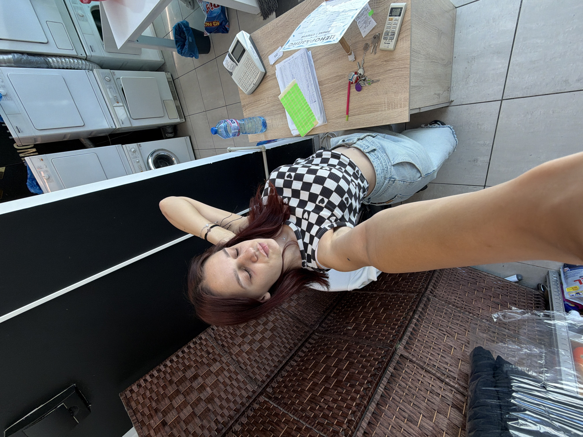 The image depicts a young woman in a laundry mat in Sofia, Bulgaria. She is wearing a black and white checkered crop top with heart cutouts and light-wash jeans. Her right arm is extended towards the camera, holding it for a selfie. Her eyes are closed, and her head is tilted back. In the background, several stacked white Miele washing machines and dryers are visible. A wooden desk is to the right, holding a calculator, papers, a bottle of water, pens, and keys. The floor is tiled.