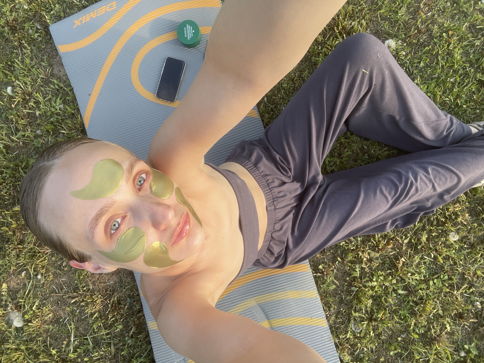 A woman is lying on a grey yoga mat with yellow stripes outdoors on grass. She is wearing a grey sports bra and grey sweatpants. She has gold eye patches on her face. A black cell phone and a green jar are on the yoga mat beside her. The location of Подшивалово, Russia, cannot be determined from the image.