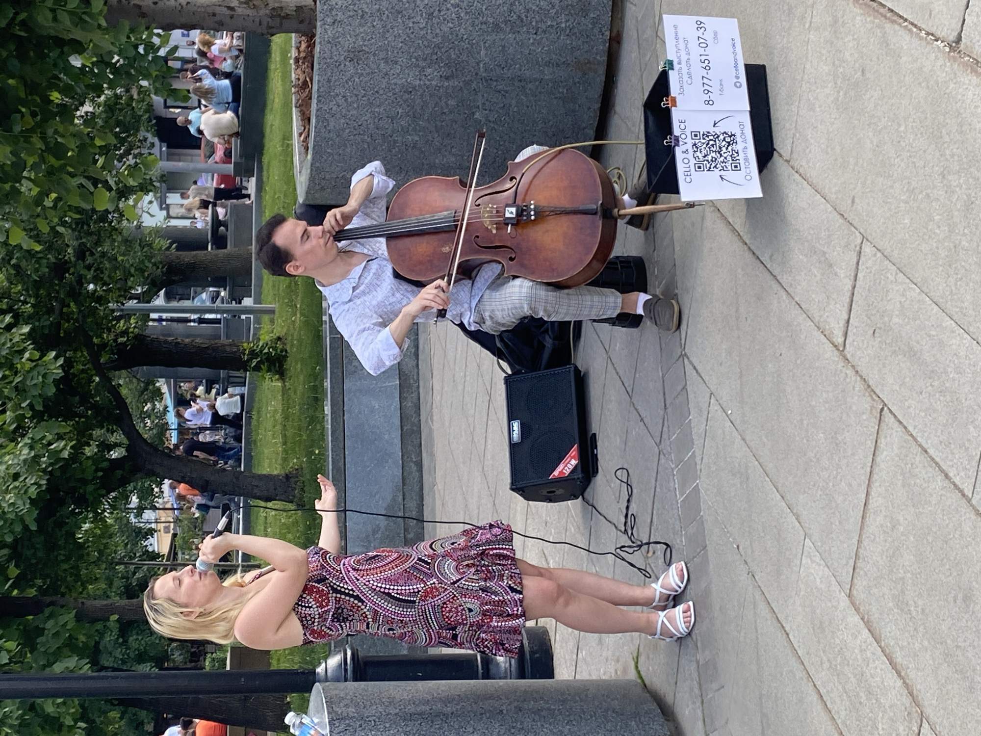 The image depicts a street performance duo in Moscow, Russia. A man plays the cello while seated, and a woman sings into a microphone. A small amplifier provides sound amplification. A sign next to the cellist invites donations via a QR code and lists contact information. There are people in the background.