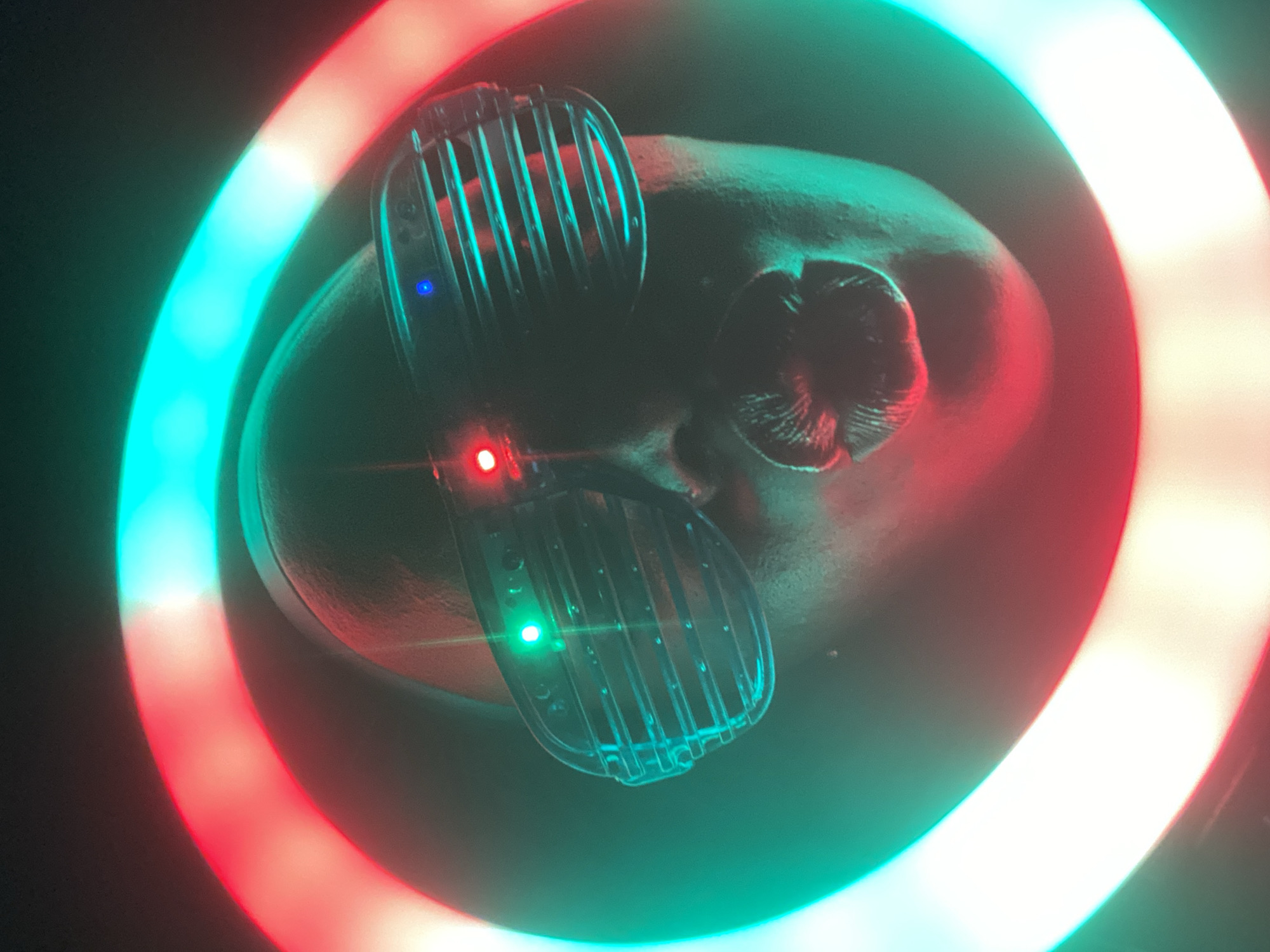 An overhead shot depicts a bald, light-skinned person wearing grill-like eyeglasses illuminated by LED lights. One lens of the glasses emits a blue light, while the other emits a red light. There is a red light and a green light in the same eyeglass lenses. The person is encircled by a large ring light that emits alternating red and green light. The scene is dark, with the lights providing the primary illumination. The location is unknown and there is no way to tell from this image.