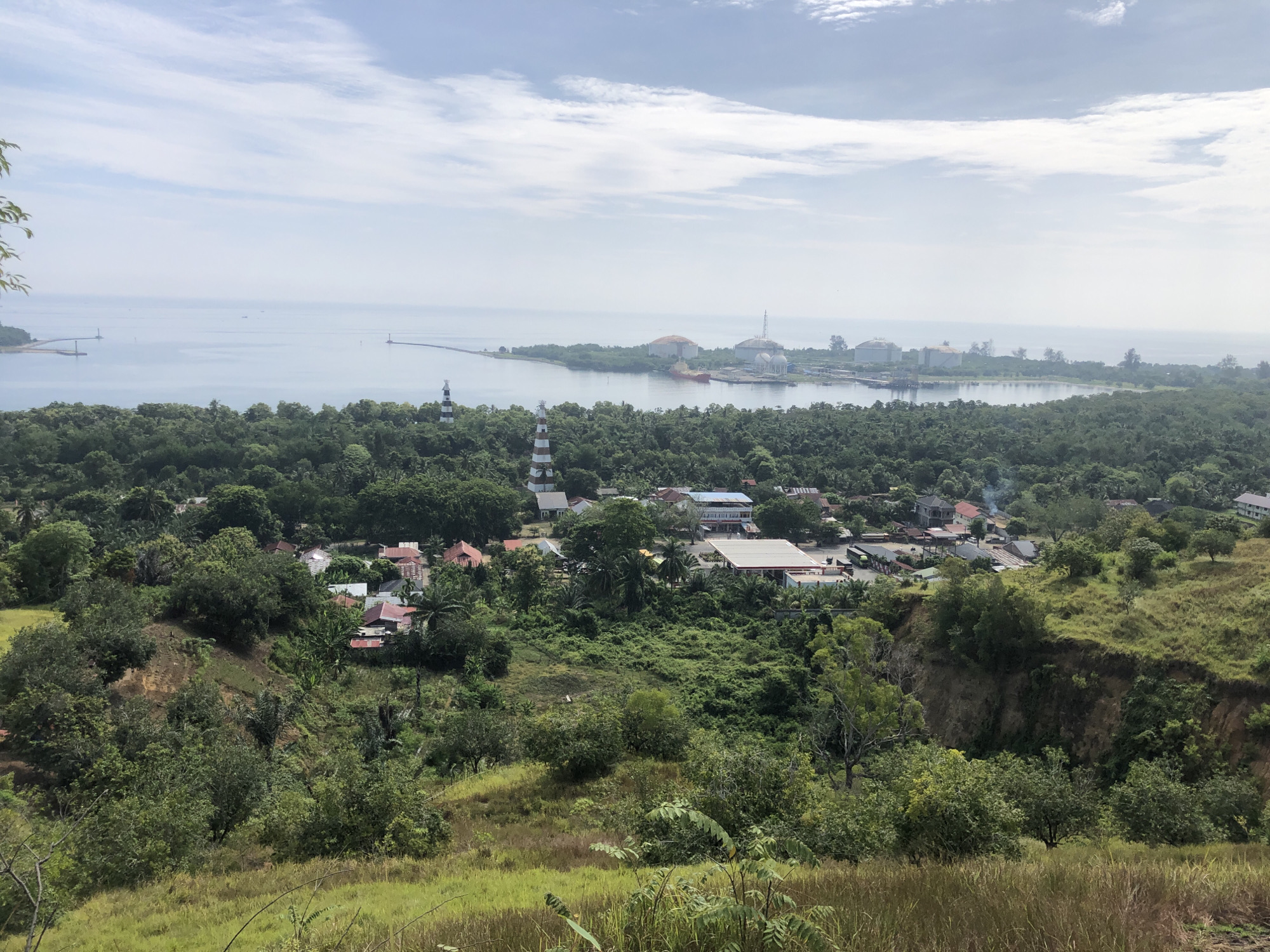 The image is an elevated, wide-angle landscape shot of a coastal area in Lhokseumawe, Indonesia, likely taken on a clear day. In the foreground, a grassy slope descends into a wooded ravine. Beyond this, a densely forested area covers rolling hills, interspersed with scattered buildings with red and white roofs, suggesting a small town or village. Two black and white striped lighthouses are visible in the mid-ground, one closer and taller than the other. Further in the distance, a body of calm, blue water stretches to the horizon. Along the coast, a long pier extends into the sea, and an industrial complex with large, spherical storage tanks and a red cargo ship is situated on the water's edge. The sky is bright with scattered white clouds. No people are visible in the image.