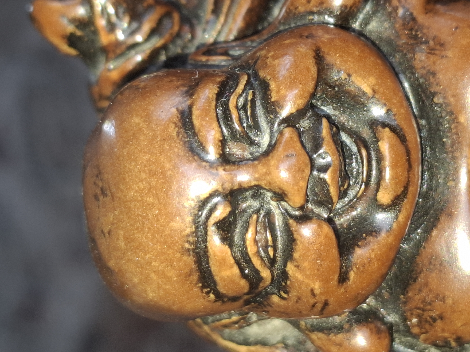 A close-up, eye-level, medium shot shows the head and upper torso of a small, carved wooden statue of a laughing Buddha. The statue is a warm, reddish-brown color with dark brown or black accents defining the features, such as the eyes and the lines of the face. The Buddha's eyes are closed in a smile, with prominent laugh lines etched around them. The face is round and full, with chubby cheeks and a wide, open-mouthed grin. The statue appears to be an antique or decorative item. The background is dark and out of focus, providing no discernible environmental context. The image was taken indoors, likely in a studio or home setting, with artificial lighting. No specific location in Almaty, Kazakhstan can be determined from this image.