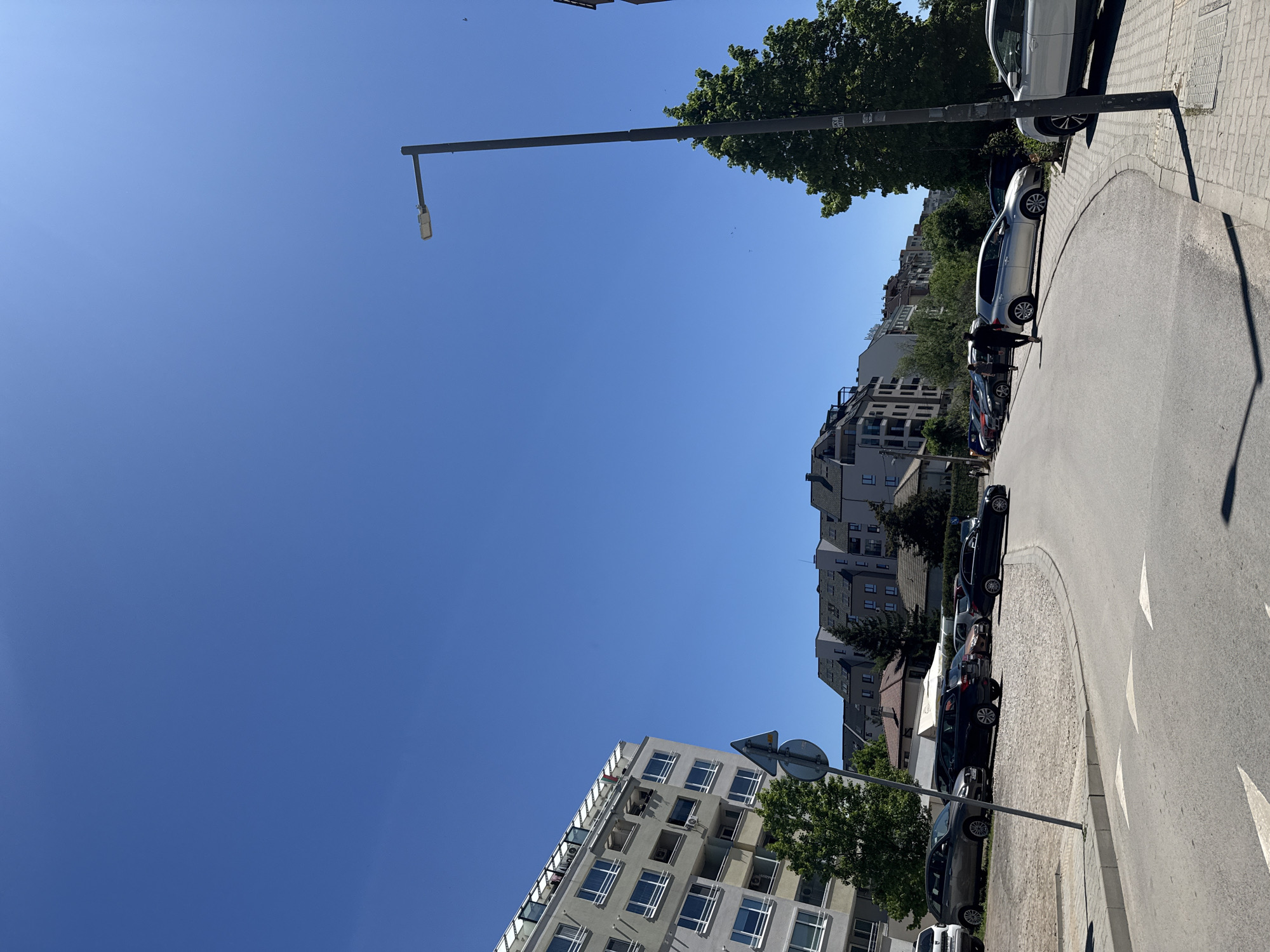 The image depicts a street scene in Sofia, Bulgaria. The sky is clear blue, and the scene is dominated by buildings, trees, parked cars along the side of the road, and a streetlight. The buildings are multistory apartment complexes with white or gray facades and multiple windows. A road is paved with grey asphalt. There are arrows painted along the street. The curb to the right of the street is lined with rectangular grey stones. Two people can be seen walking along the side of the road.